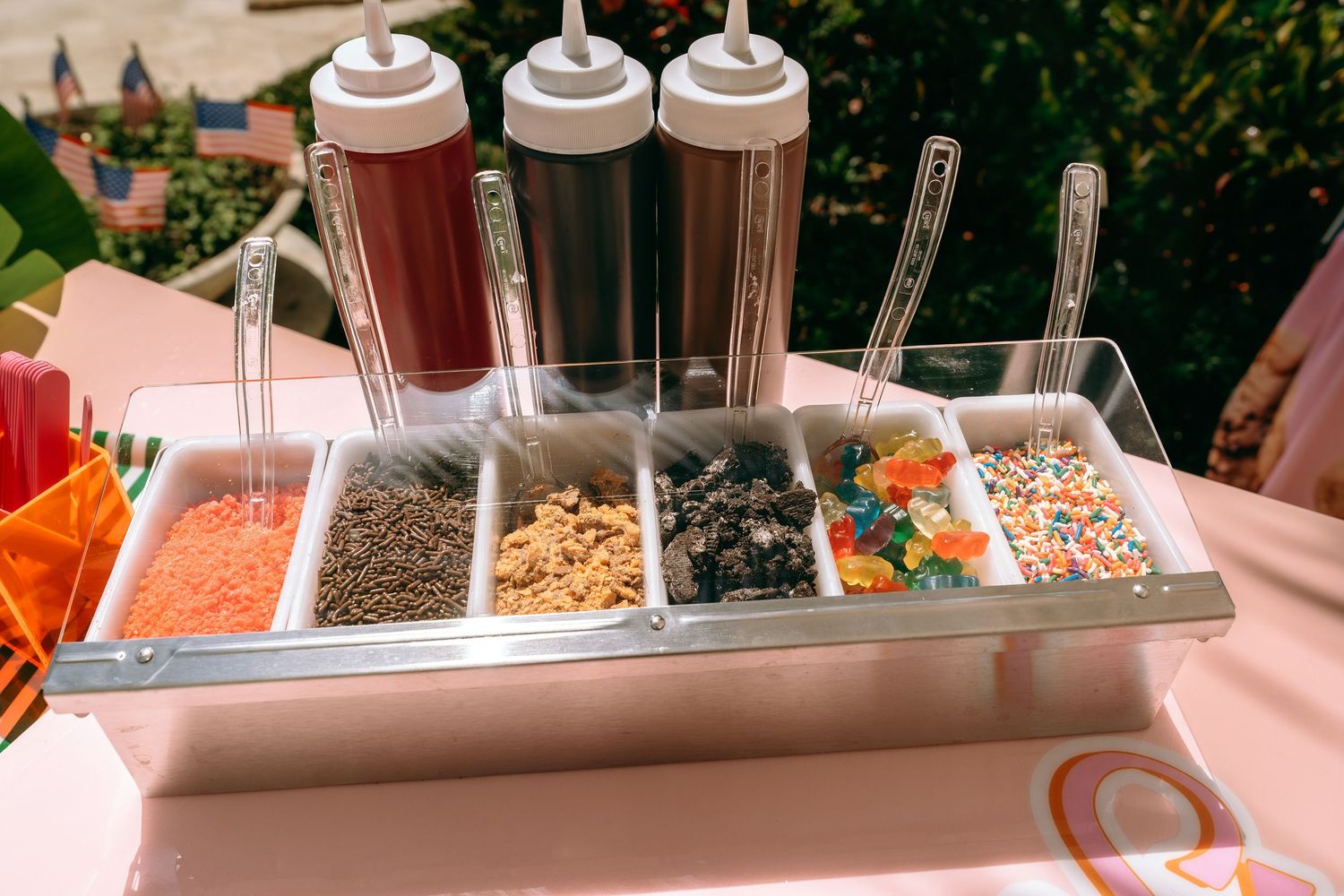 Ice cream topping bar with sprinkles, nuts, and candy in metal tray with chocolate and caramel sauce bottles.