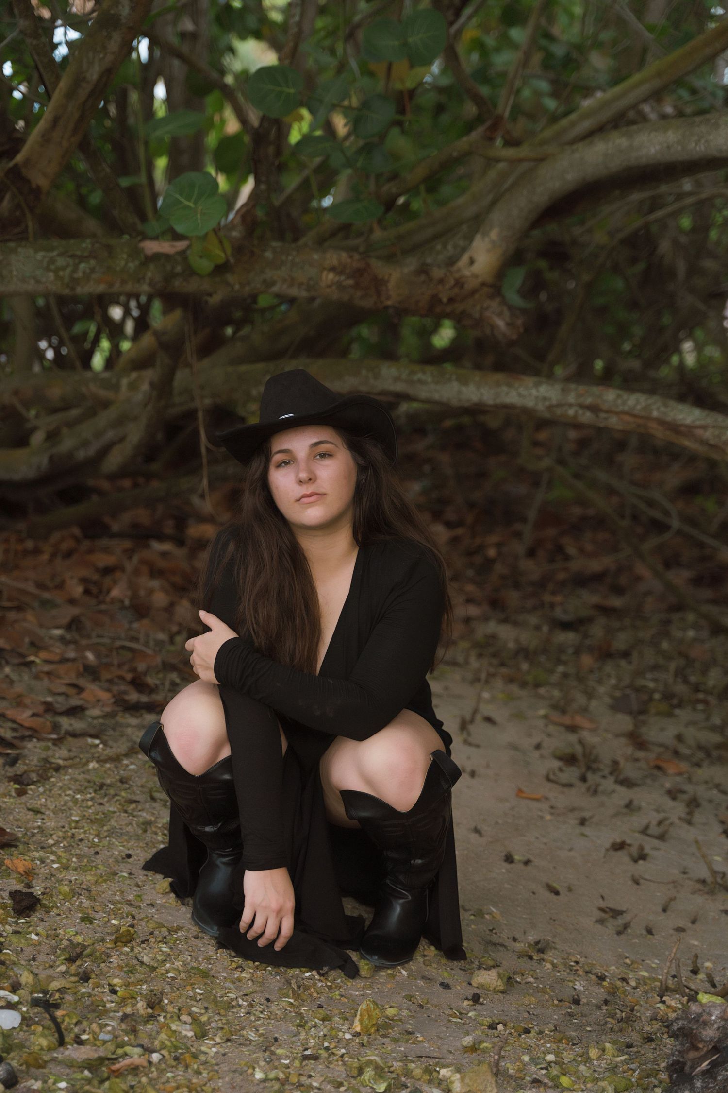 Woman in black dress and hat poses on ground beneath tree branches.