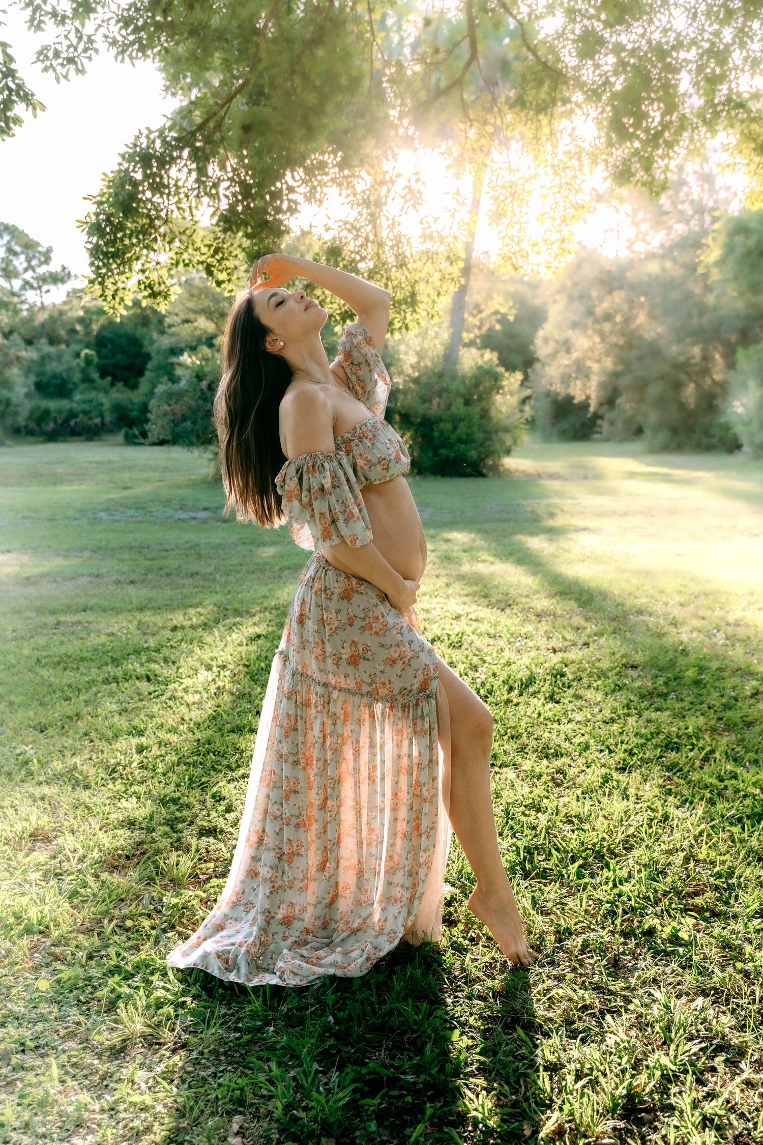 Ethereal maternity portraits in a floral dress against sun-drenched grass and trees.