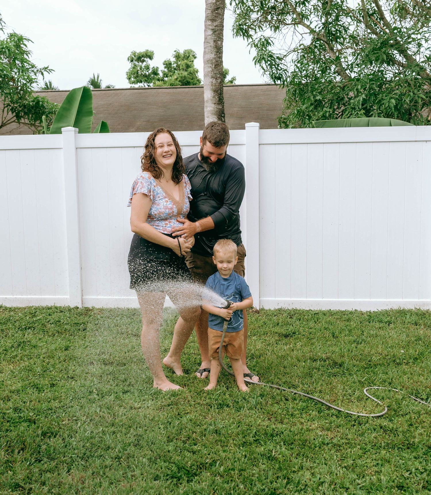 Family of three playing with garden hose on green lawn, toddler spraying water, white fence background.