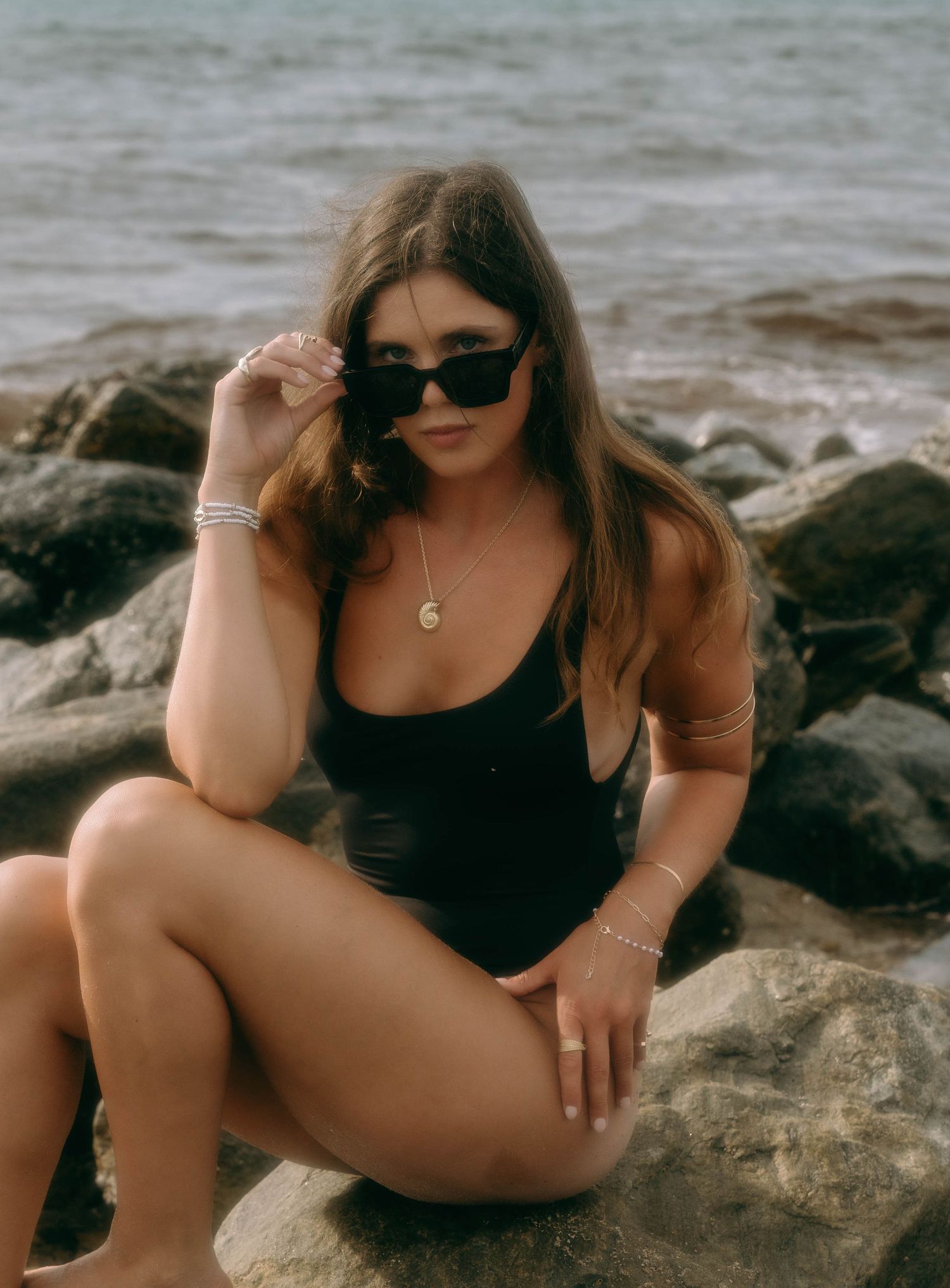 Woman in black swimsuit and sunglasses sitting on rocks by the ocean shore.