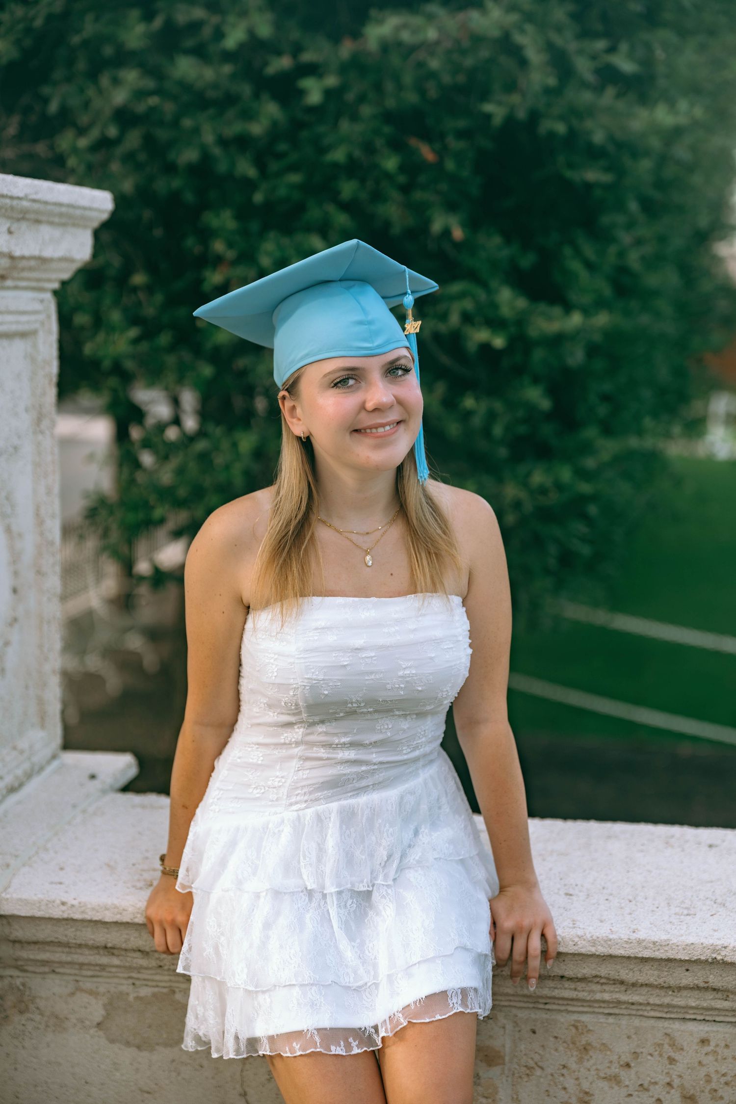 Young woman in white dress and light blue graduation cap smiling outdoors near marble columns.