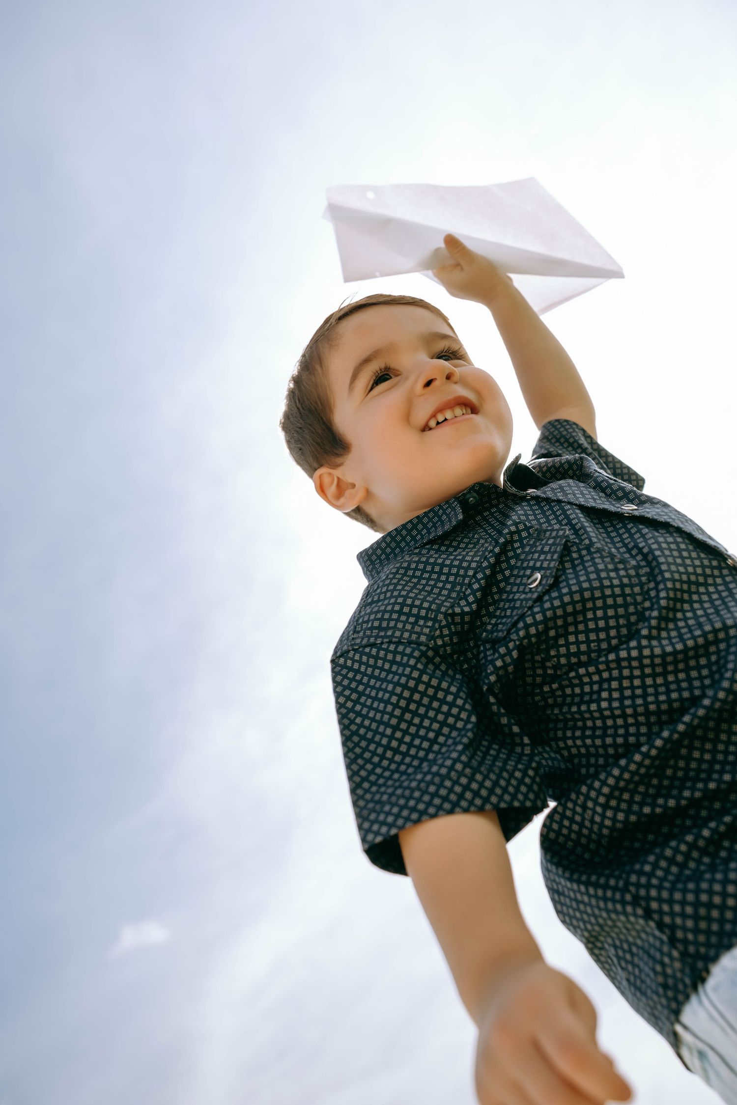 Smiling boy holding a paper airplane up in the air against a bright sky.