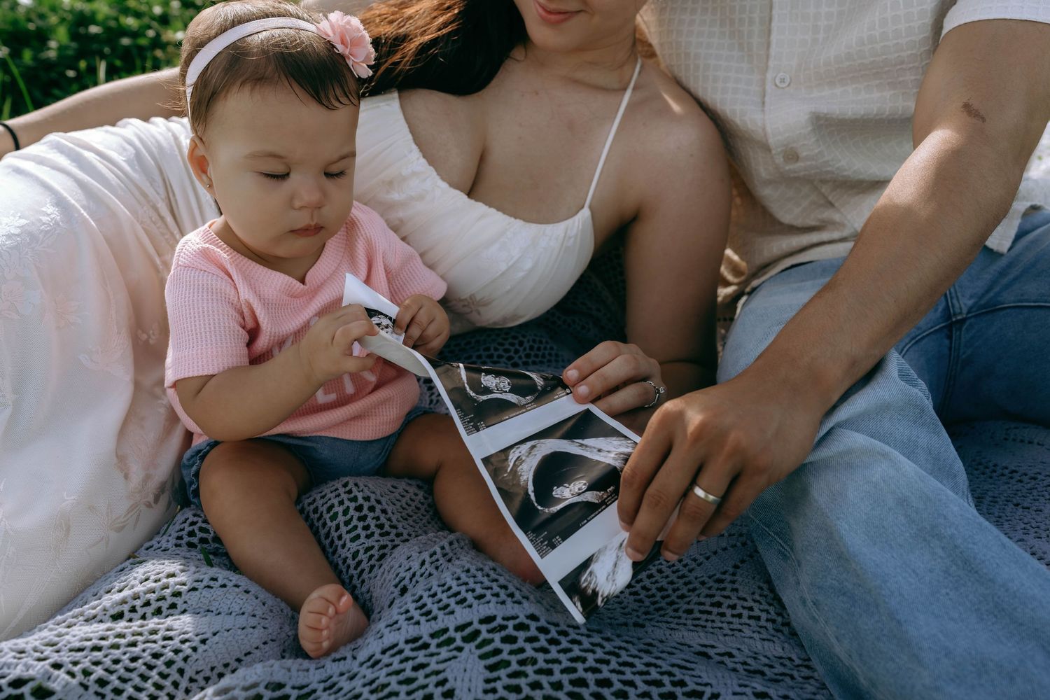 A couple sits outdoors with their baby girl in pink, looking at ultrasound photos together.