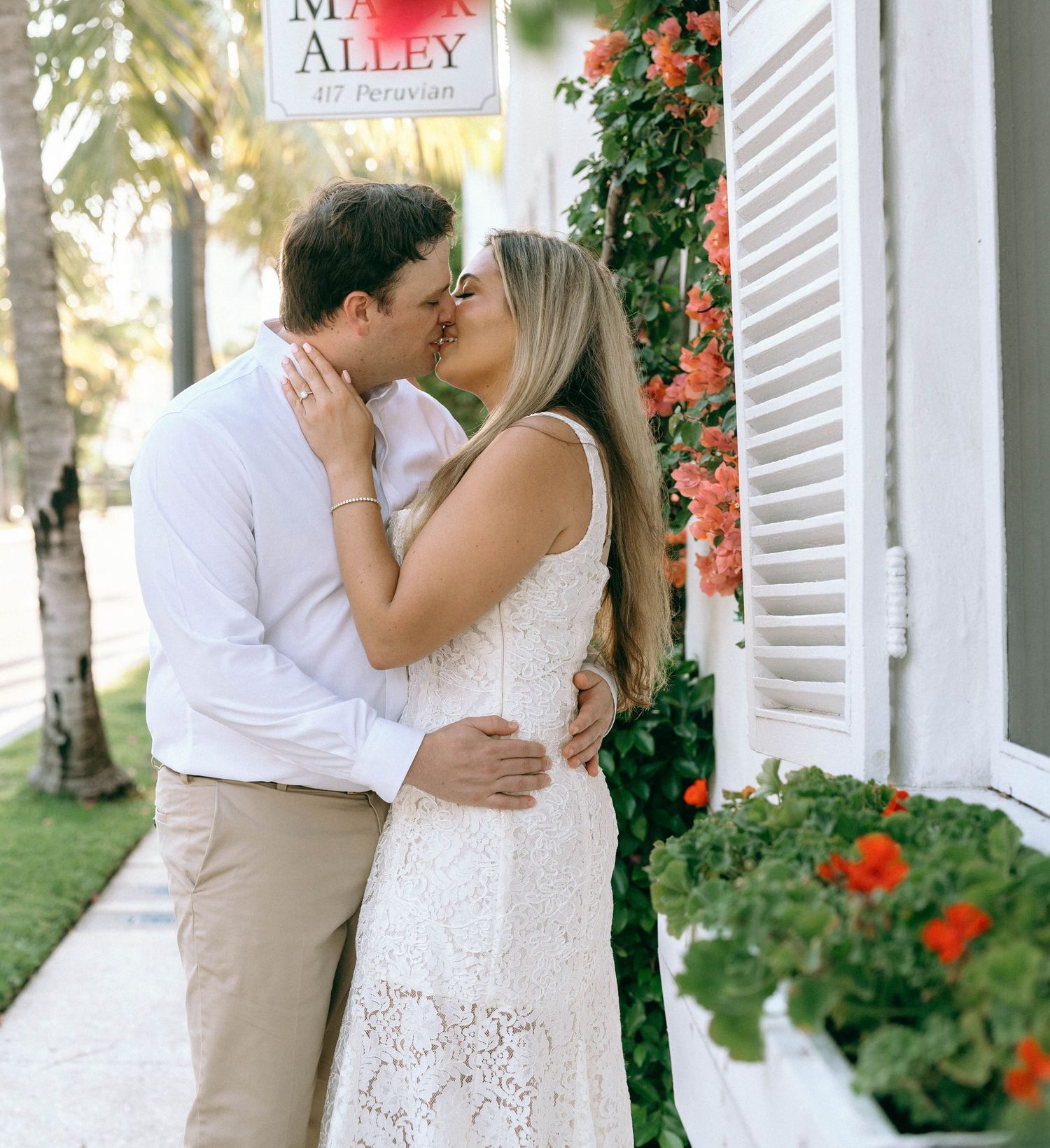 Couple sharing a romantic kiss outside a white building with red flowers, woman in white lace dress.