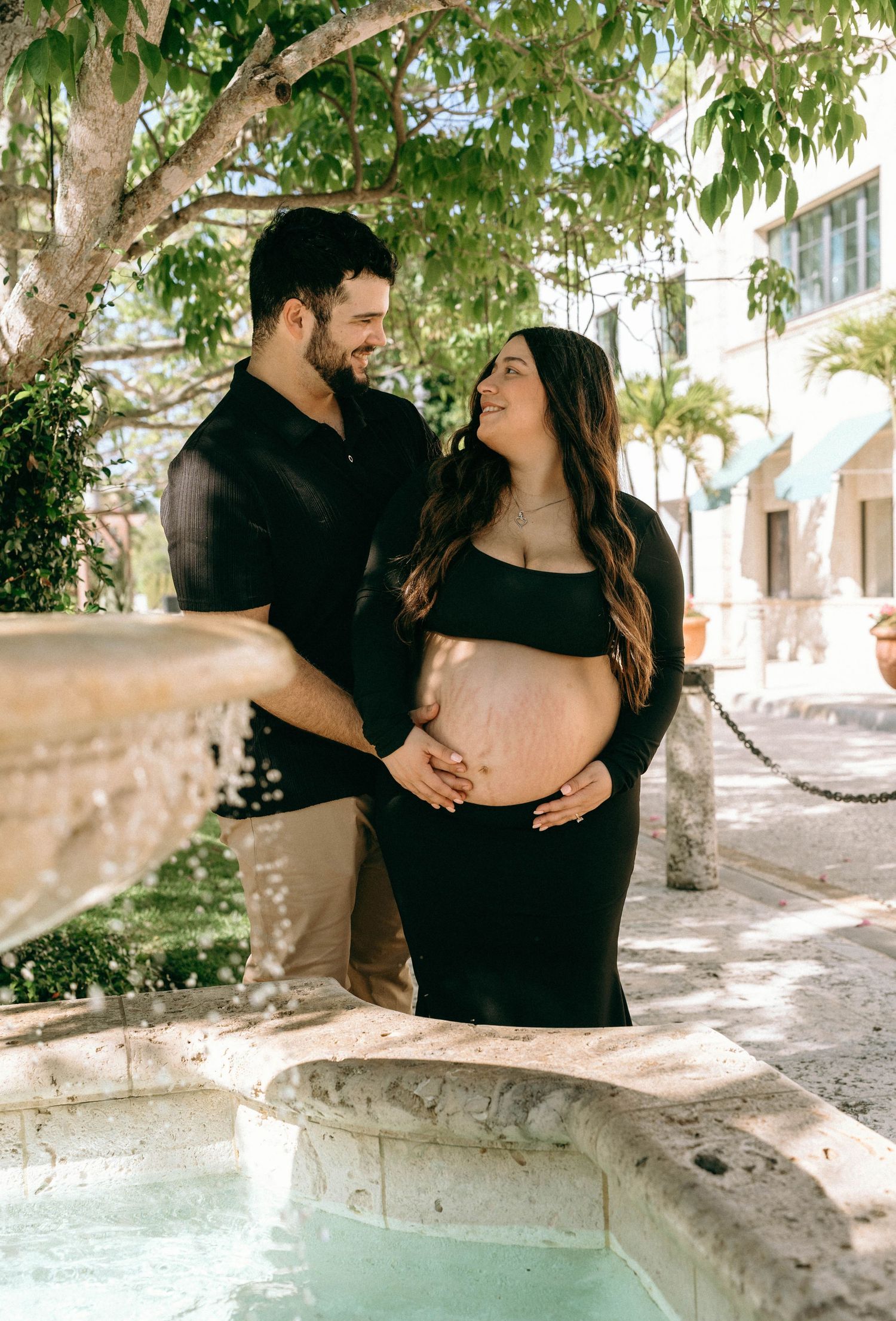 Couple sharing a tender moment by a fountain, cradling pregnant belly in matching black outfits outdoors. Worth Avenue, Palm Beach, FL