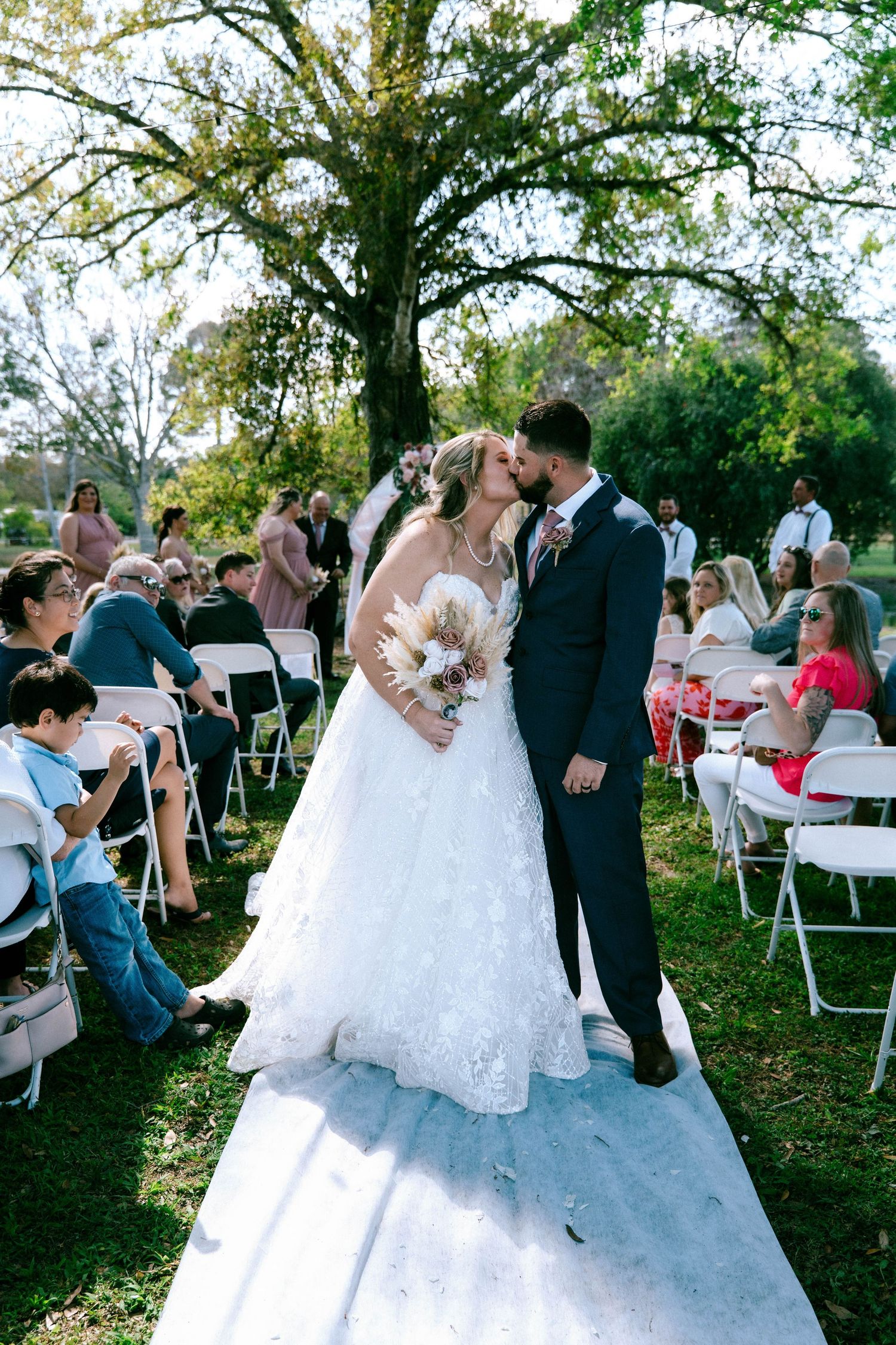 Bride and groom share first kiss as newlyweds walking down outdoor wedding aisle surrounded by seated guests.