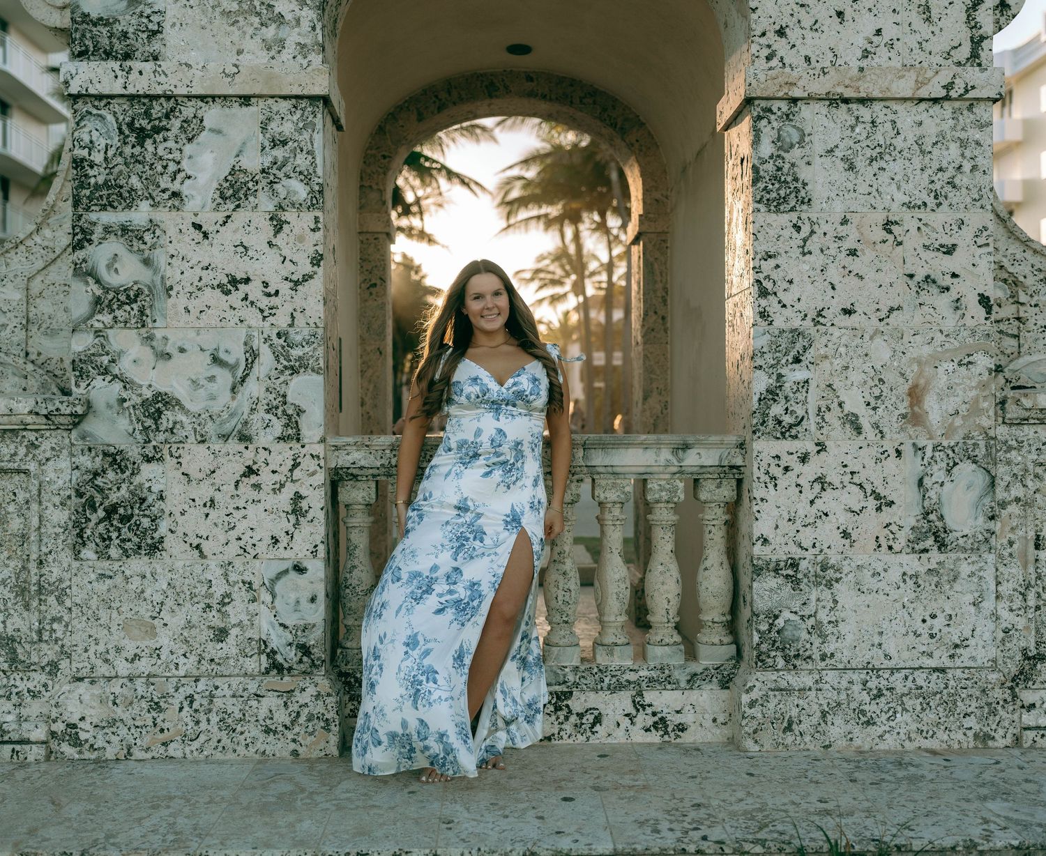 Woman in white blue floral slit maxi dress posing under stone archway at sunset.