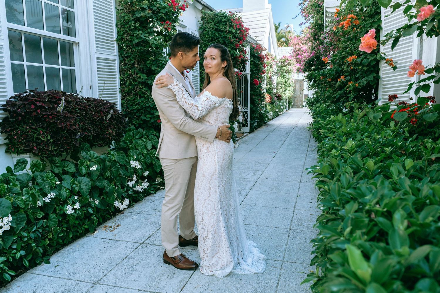 Couple embracing in wedding attire surrounded by lush greenery and roses on a garden pathway.