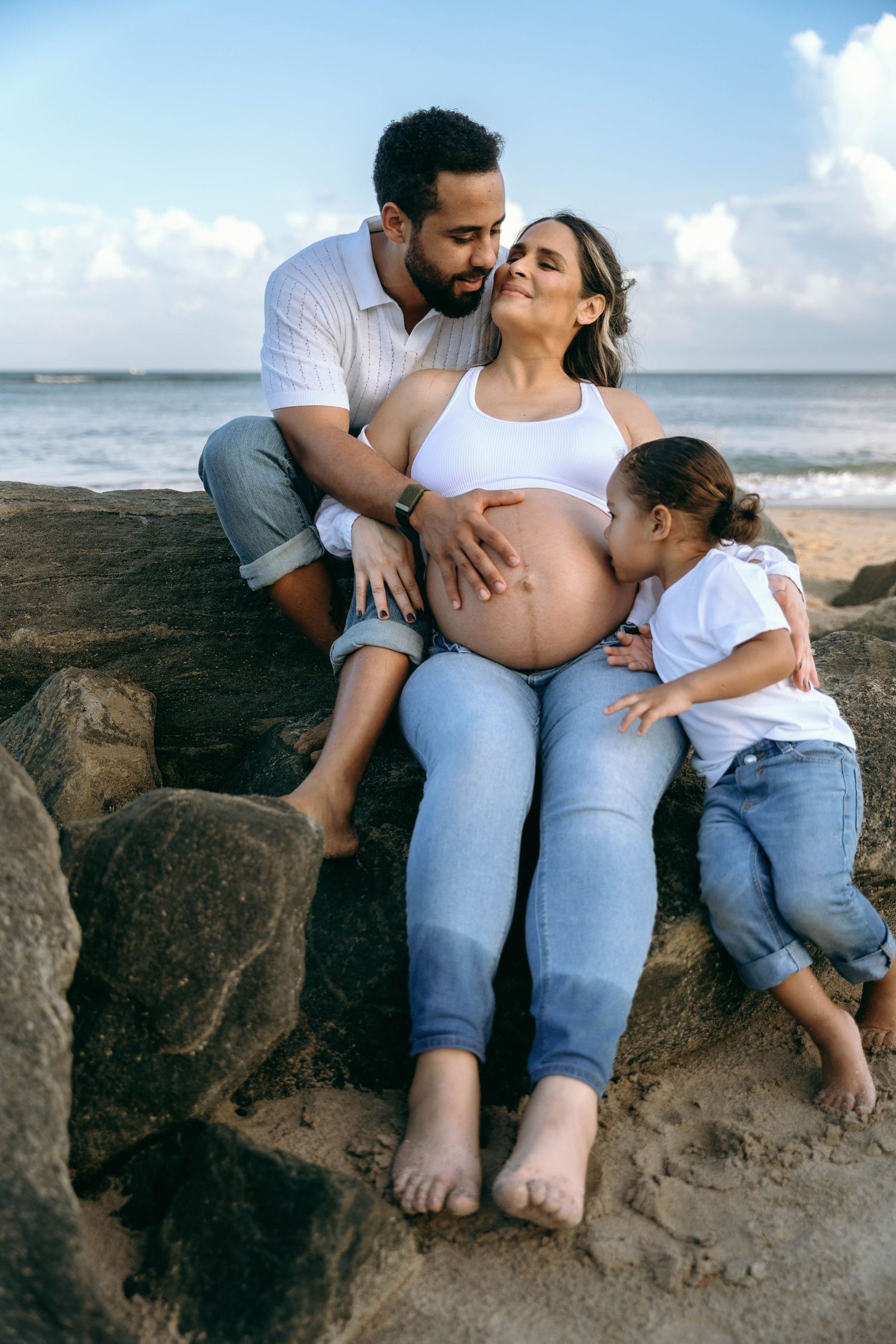 Family sits together on rocky beach formation during sunset maternity session.