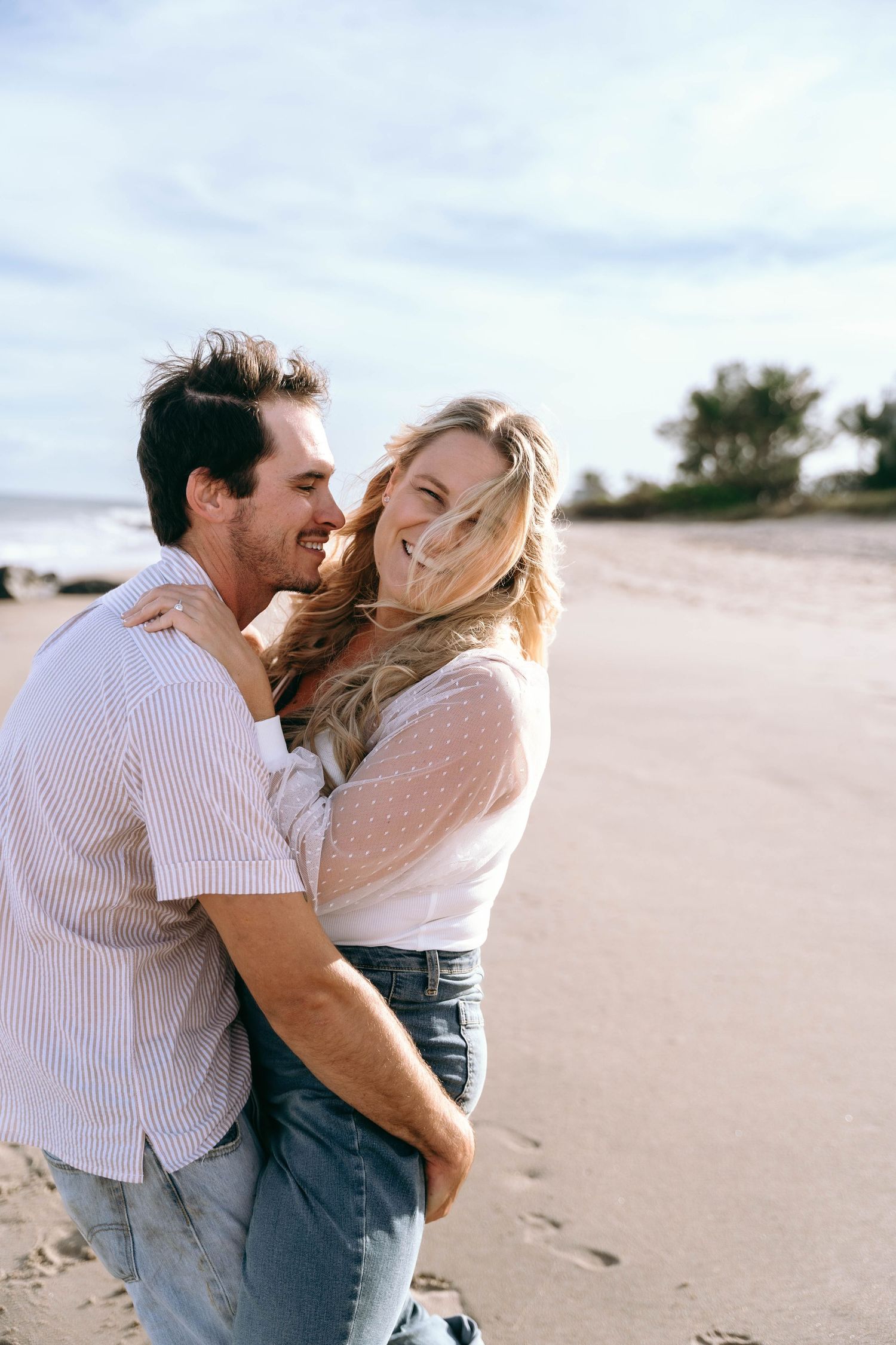 Romantic couple embraces and laughs together on a sandy beach during sunset.