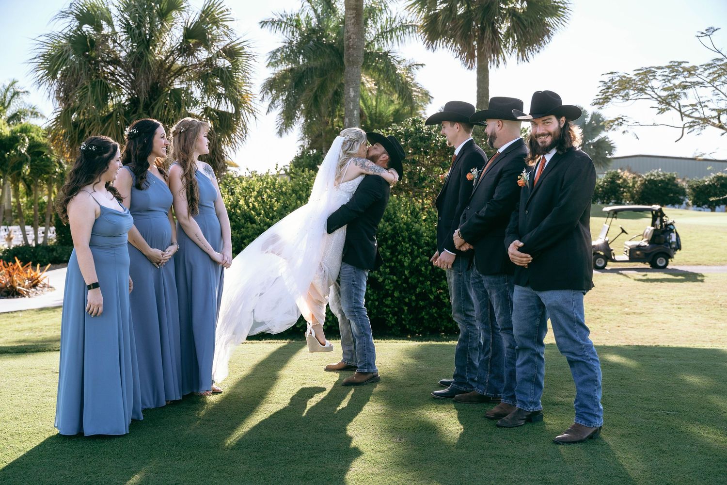 Wedding party in blue dresses and cowboy attire gather on a palm-lined lawn for photos.