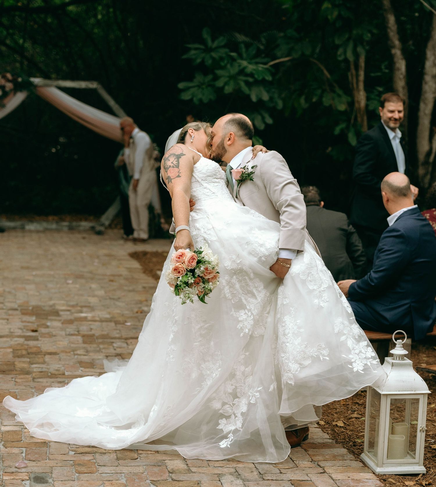 A romantic wedding kiss on a brick patio with decorative lanterns and floral bouquet in a garden setting.
