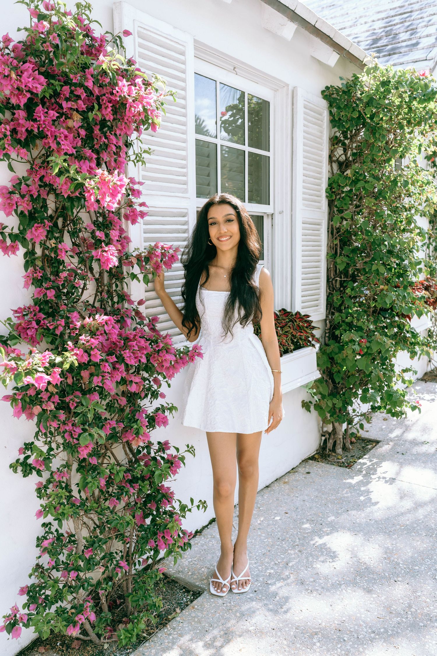 Young woman in white mini dress posing by pink bougainvillea flowers near white building on Worth Avenue Palm beach.