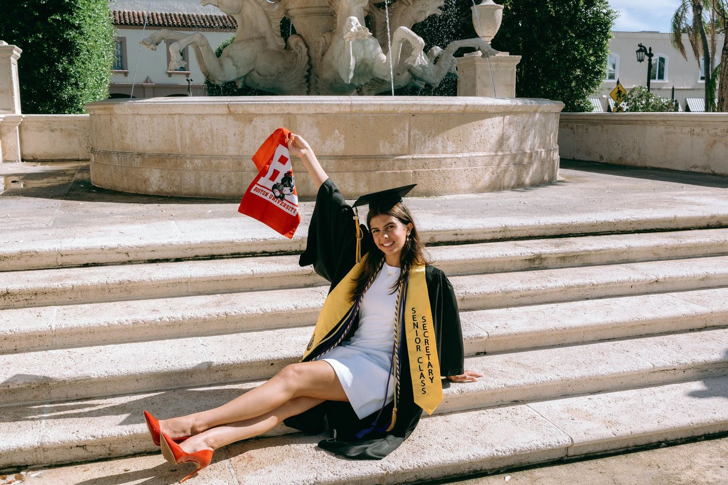 Graduate in cap and gown sitting on fountain steps holding Boston University pennant on Worth Avenue Palm beach.