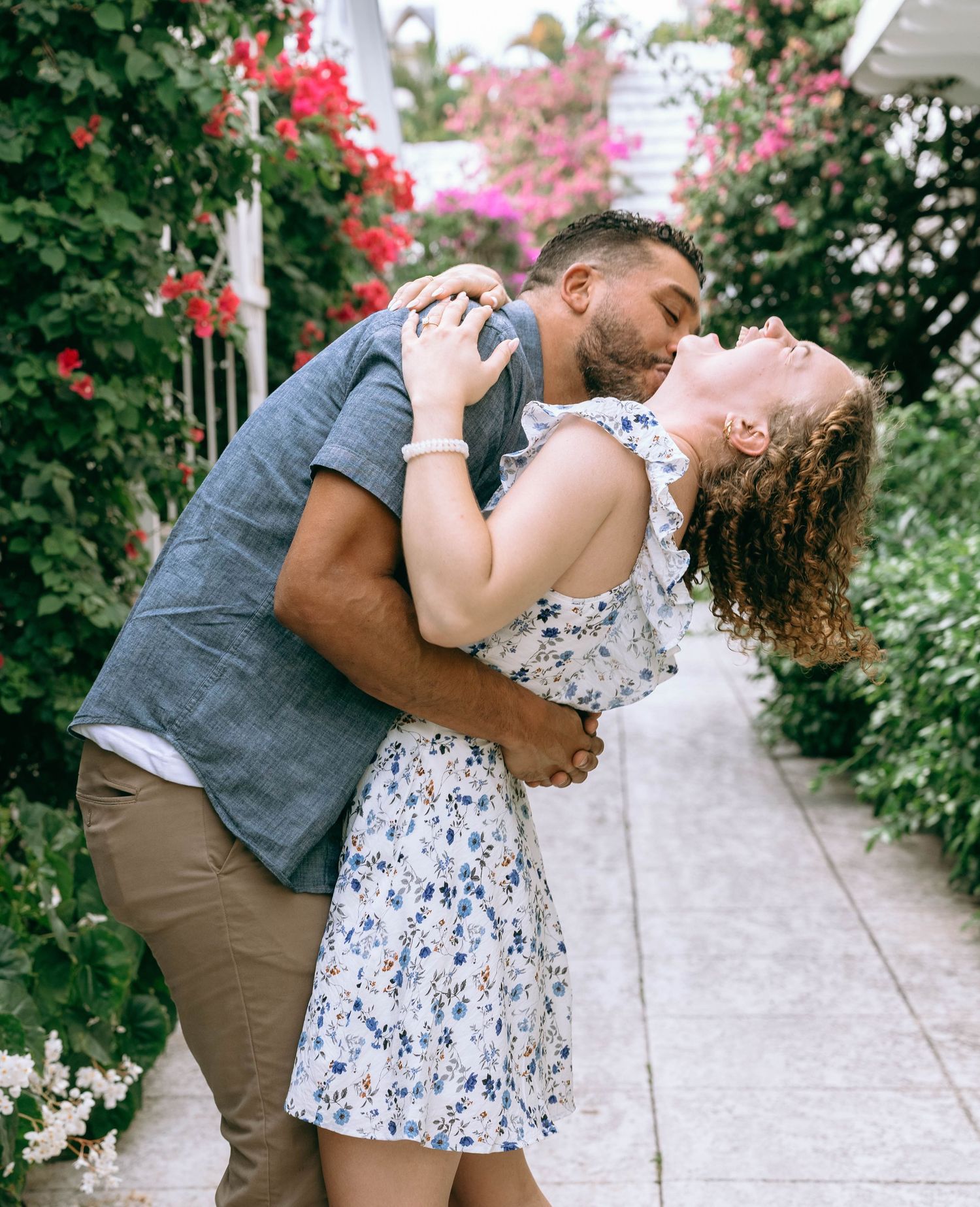 A couple shares a romantic kiss in a flower-lined garden path, the woman in a floral dress leaning back joyfully Engagement photos on Worth