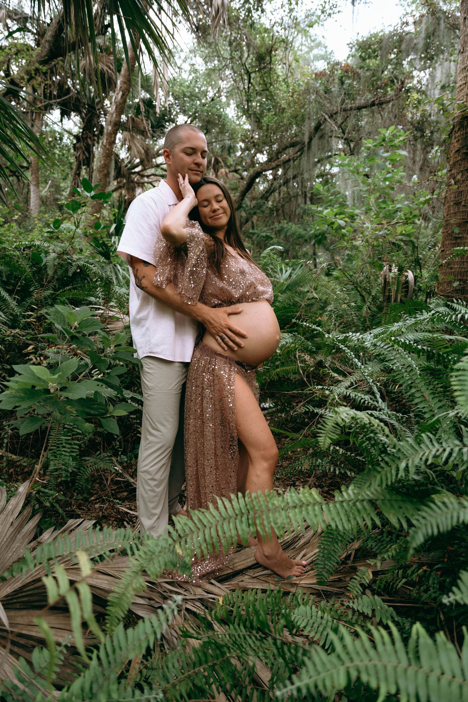 Couple embraces among lush ferns during outdoor maternity photography session.