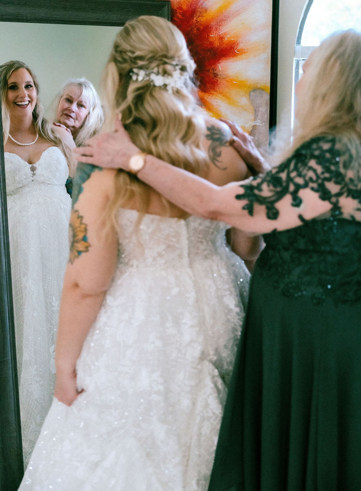 Mother helps bride in lace wedding gown prepare, reflected in mirror during bridal getting ready moment.