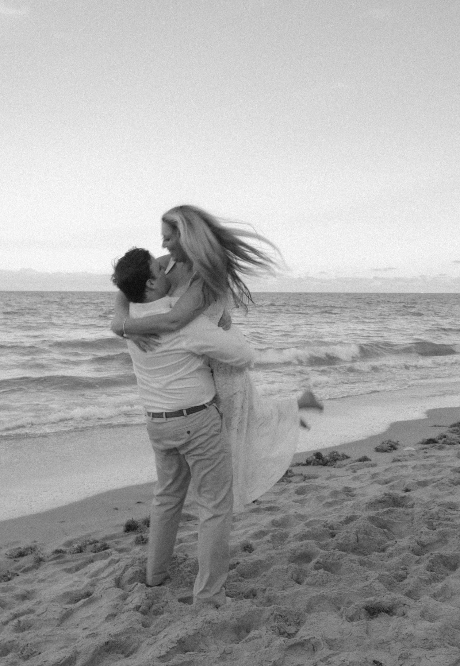 Black and white beach photo of a man lifting his partner joyfully by the ocean.