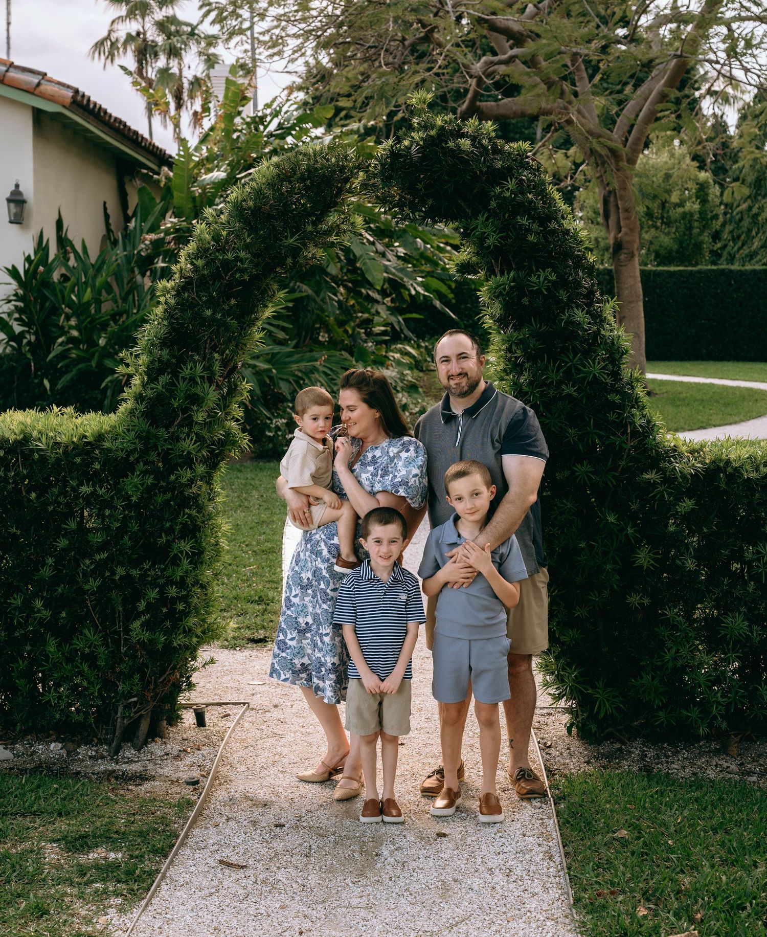 Family of five posing under a lush green garden arch on a stone pathway, dressed in coordinating blue outfits.  Family photos at Bradley