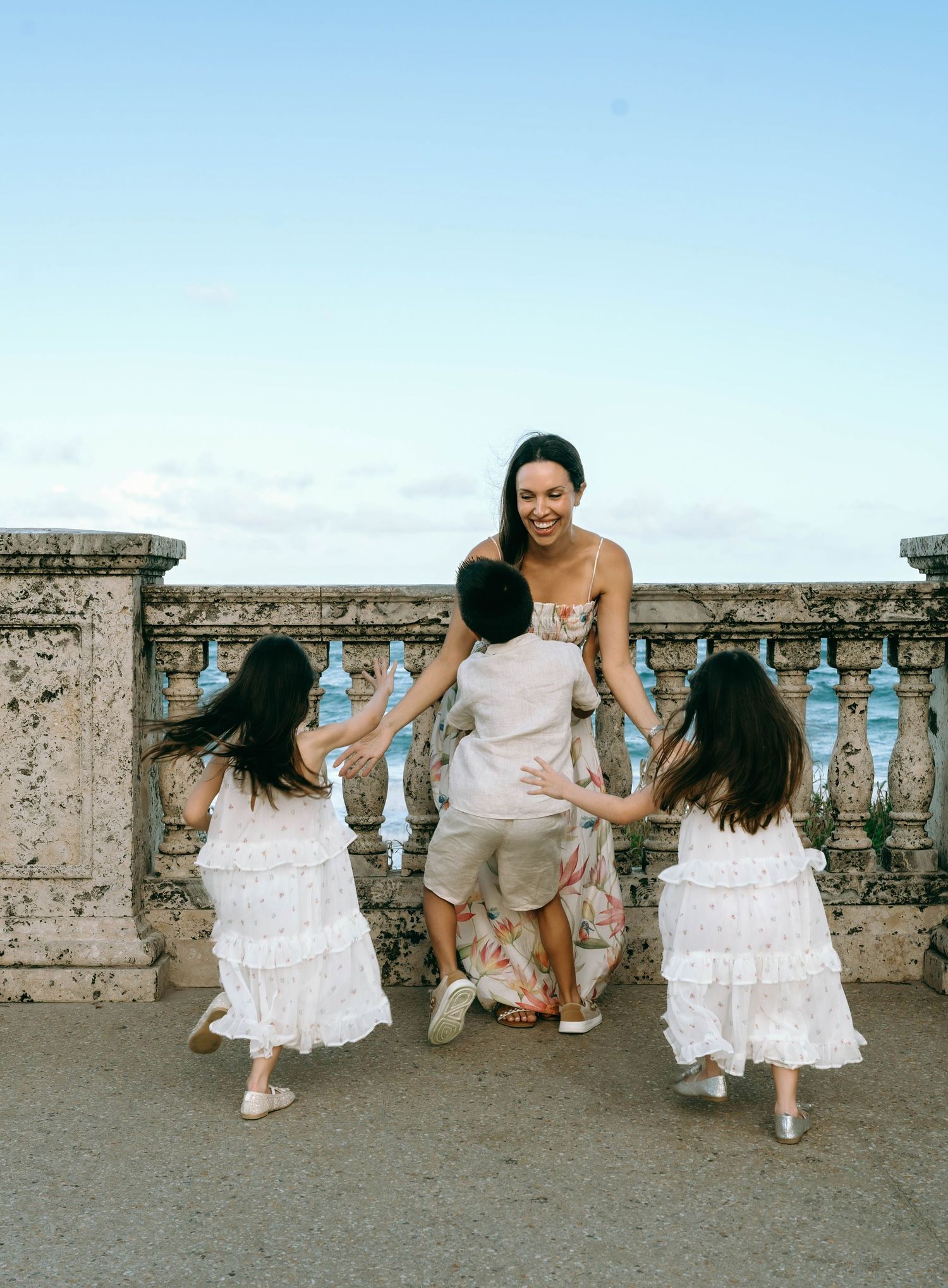 A joyful mother in a floral dress laughs as her three children run toward her on a scenic stone terrace. Worth Avenue, Palm Beach, FL