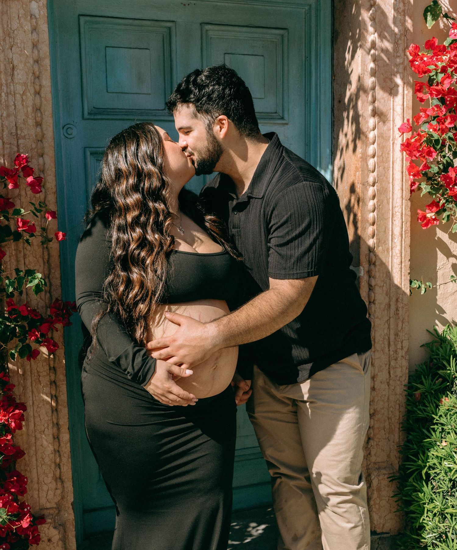 Pregnant couple kissing in front of teal door surrounded by red flowers, man cradling baby bump outdoors. Worth Avenue, Palm Beach, FL