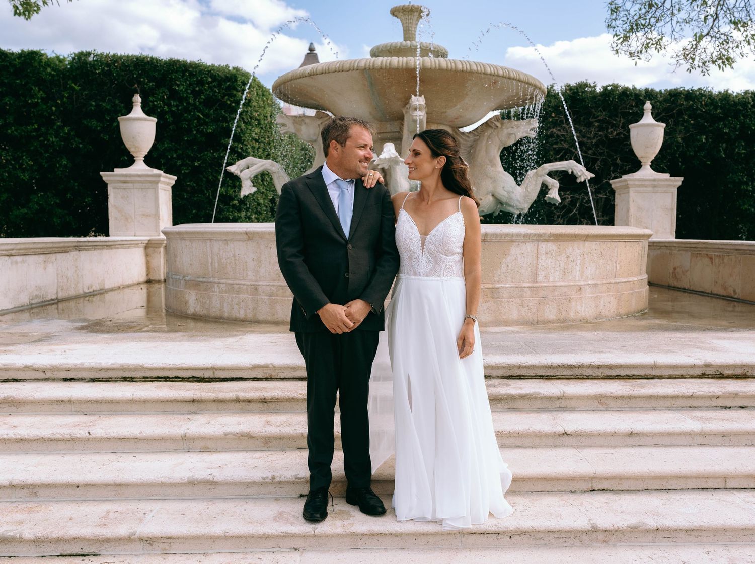 Bride and groom smiling at each other in front of an elegant stone fountain at their outdoor wedding venue.