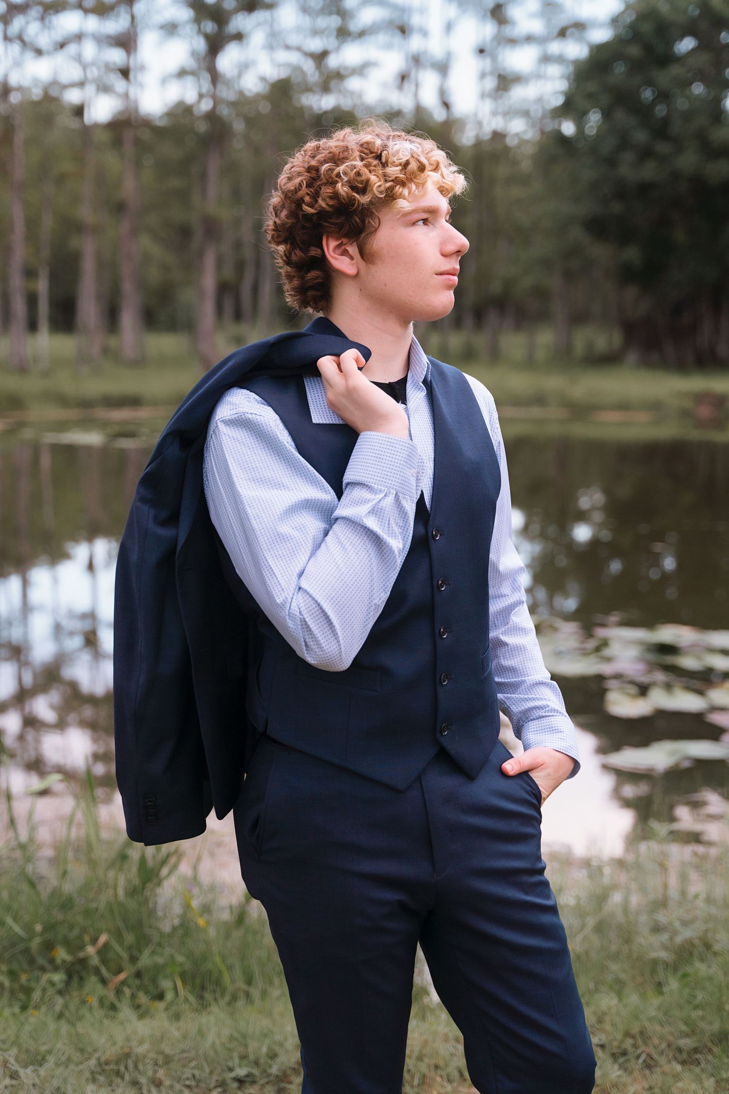 Formal portraits in a navy vest and white shirt by a scenic pond.