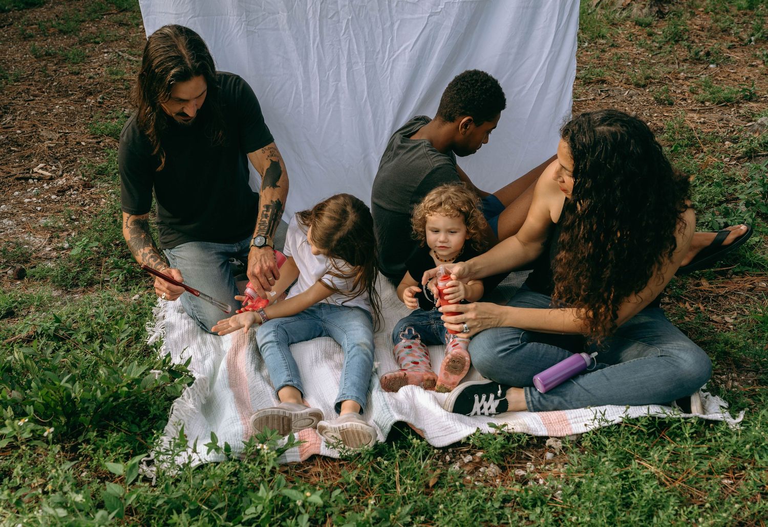 Family with two young children sitting on a blanket outdoors, playing together near a white backdrop in a grassy area.