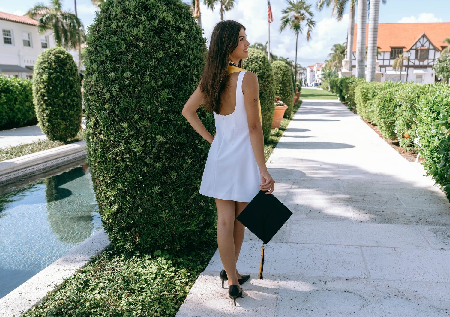 Graduate in white mini dress and heels holding cap while walking along garden pathway on Worth Avenue Palm beach.