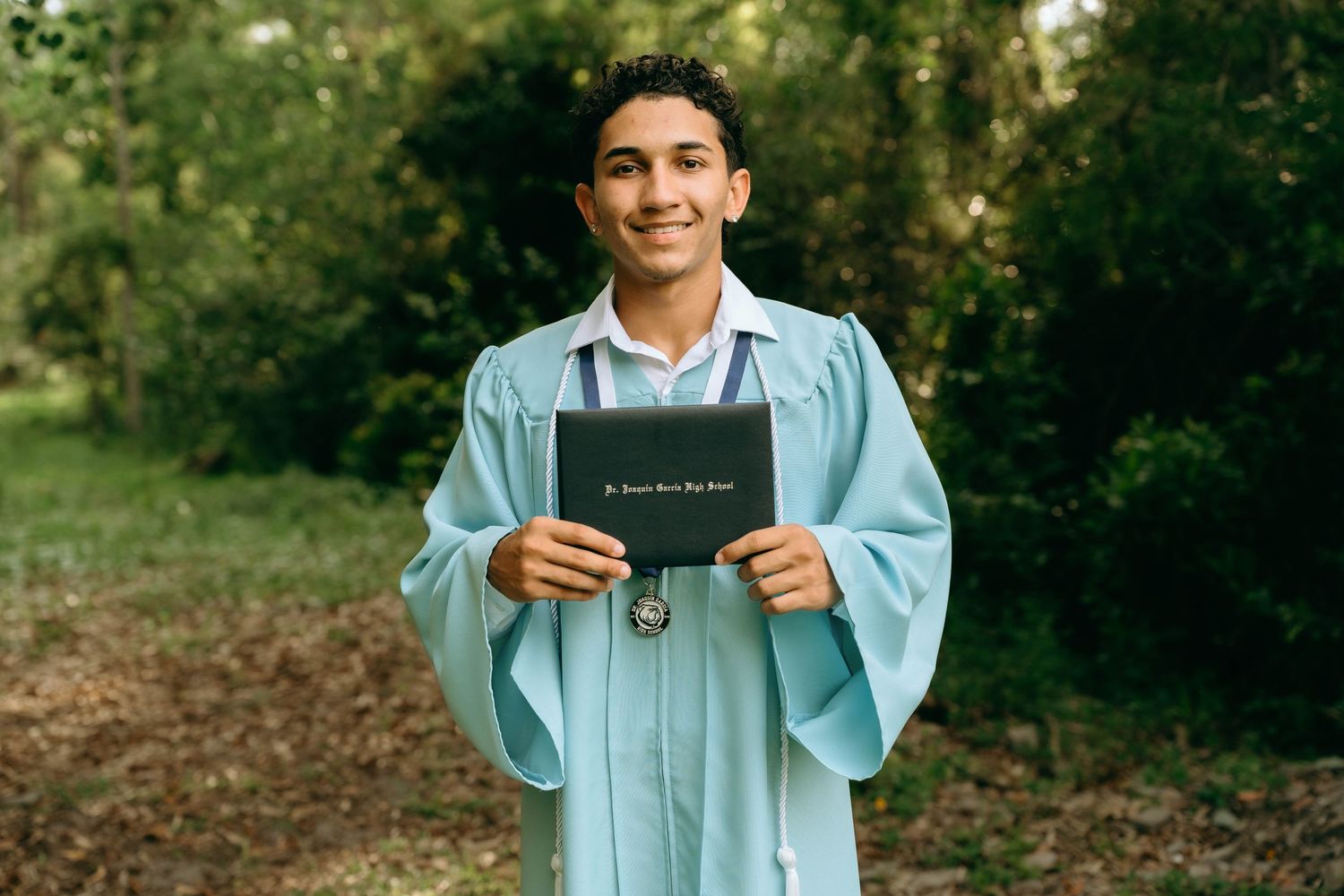 Young man in light blue graduation gown proudly holding diploma outdoors with green trees in background. Senior Photos at Colt Farms