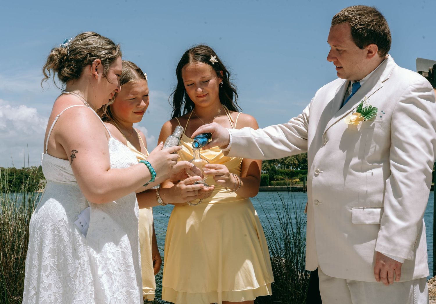 Bride and groom perform sand ceremony with guests at outdoor waterfront wedding on sunny day.
