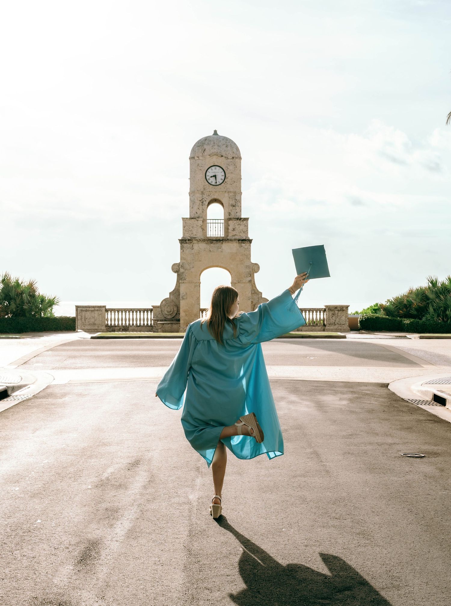 Graduate in teal cap and gown walks toward historic clock tower holding diploma on sunny day.