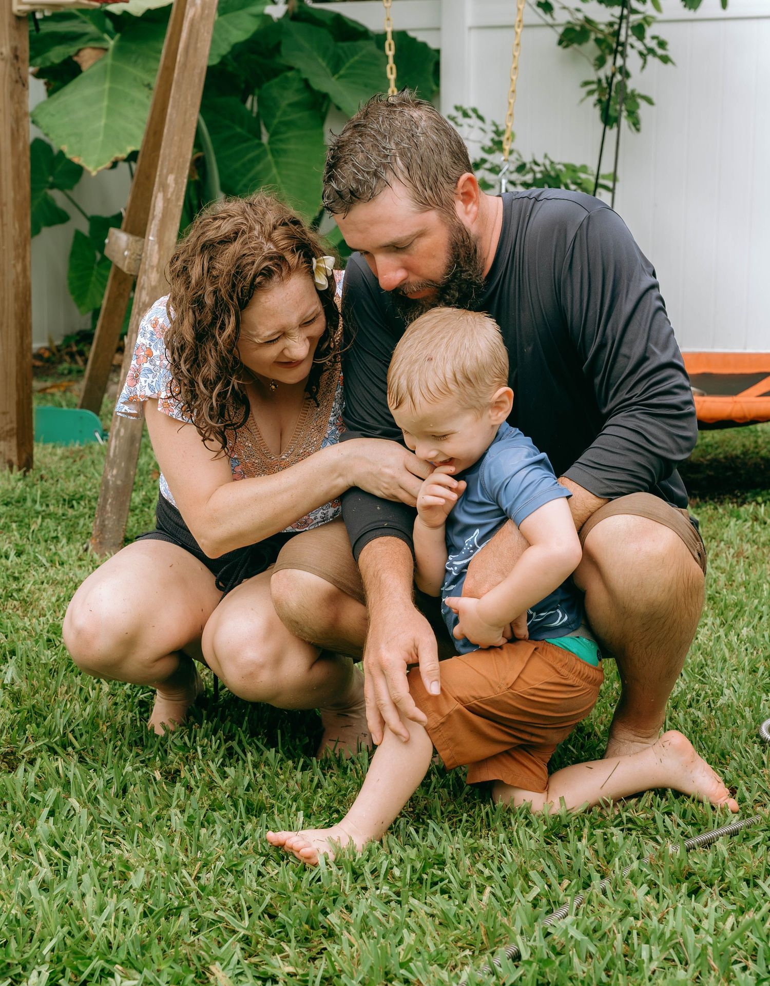 A loving family crouches on green grass, parents smiling at their young toddler boy in a sunny backyard.