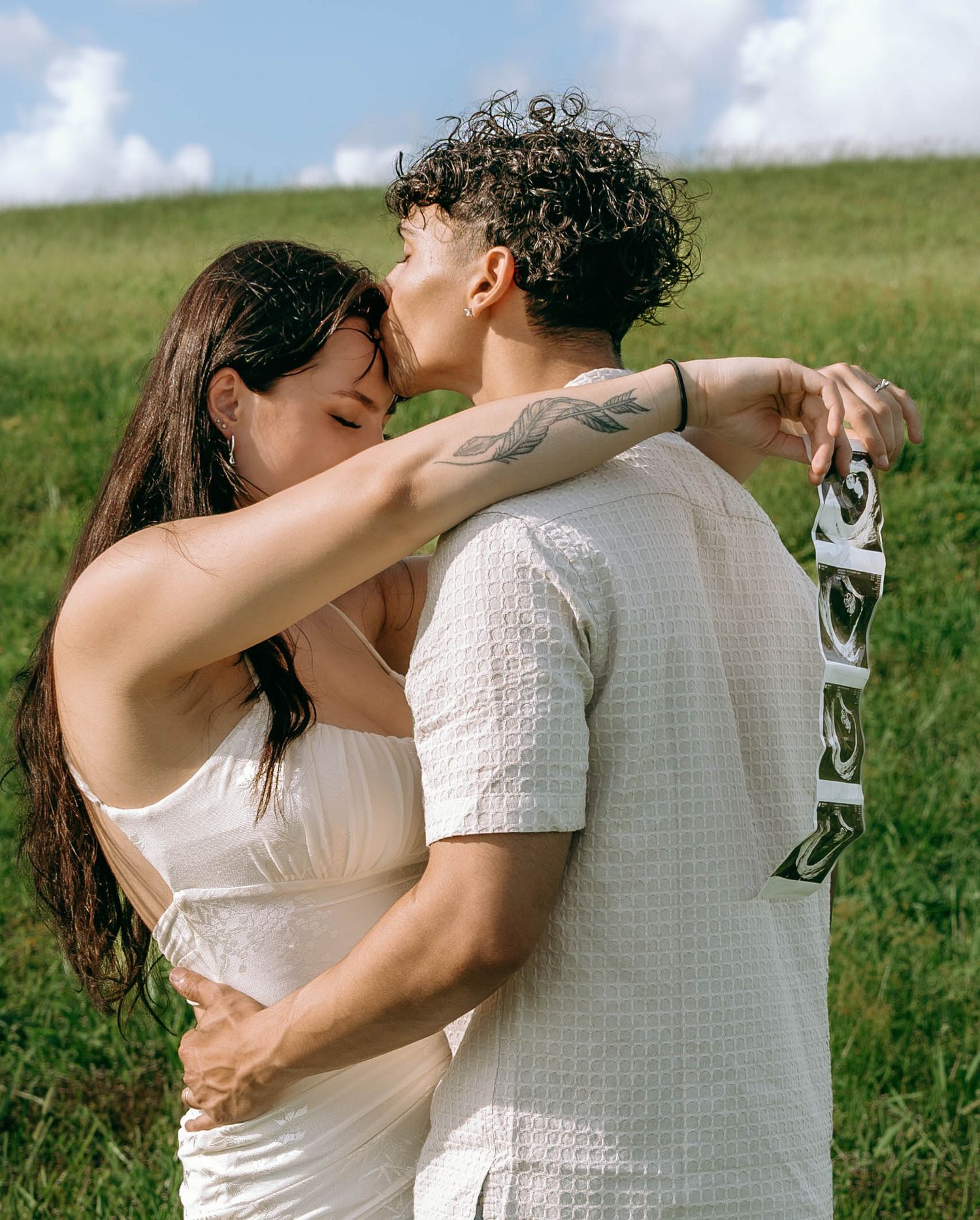Couple embracing and kissing in a green field, woman in white dress holding ultrasound photos on a sunny day.
