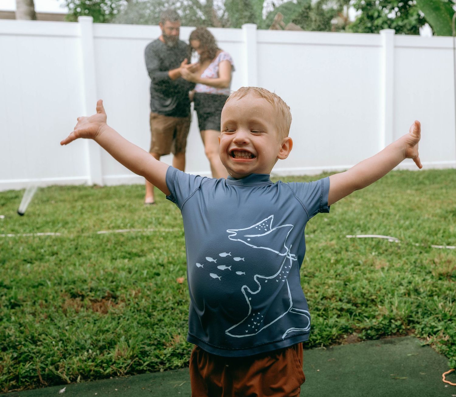 Excited toddler boy in shark t-shirt raises arms wide outdoors with parents in background.