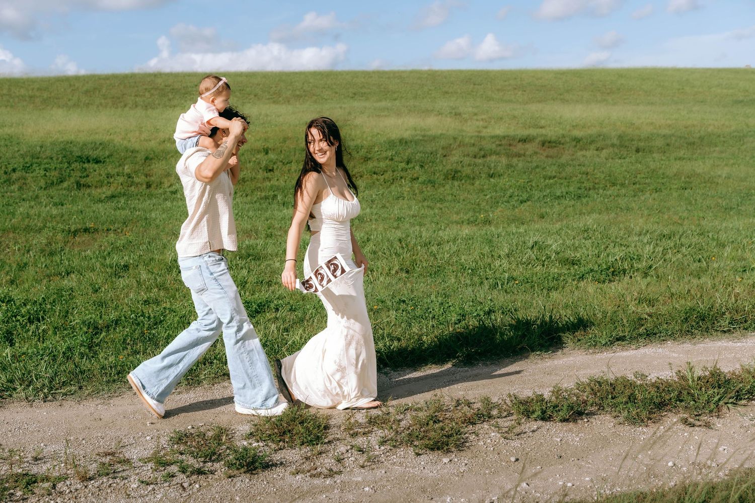 A happy family walks along a dirt path in a green field, with a woman in a white dress and a man carrying a child.