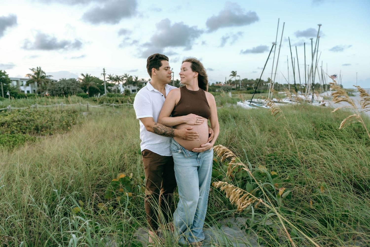 Couple embracing outdoors near marina, pregnant woman in brown top and jeans, partner in white shirt, lush greenery background.