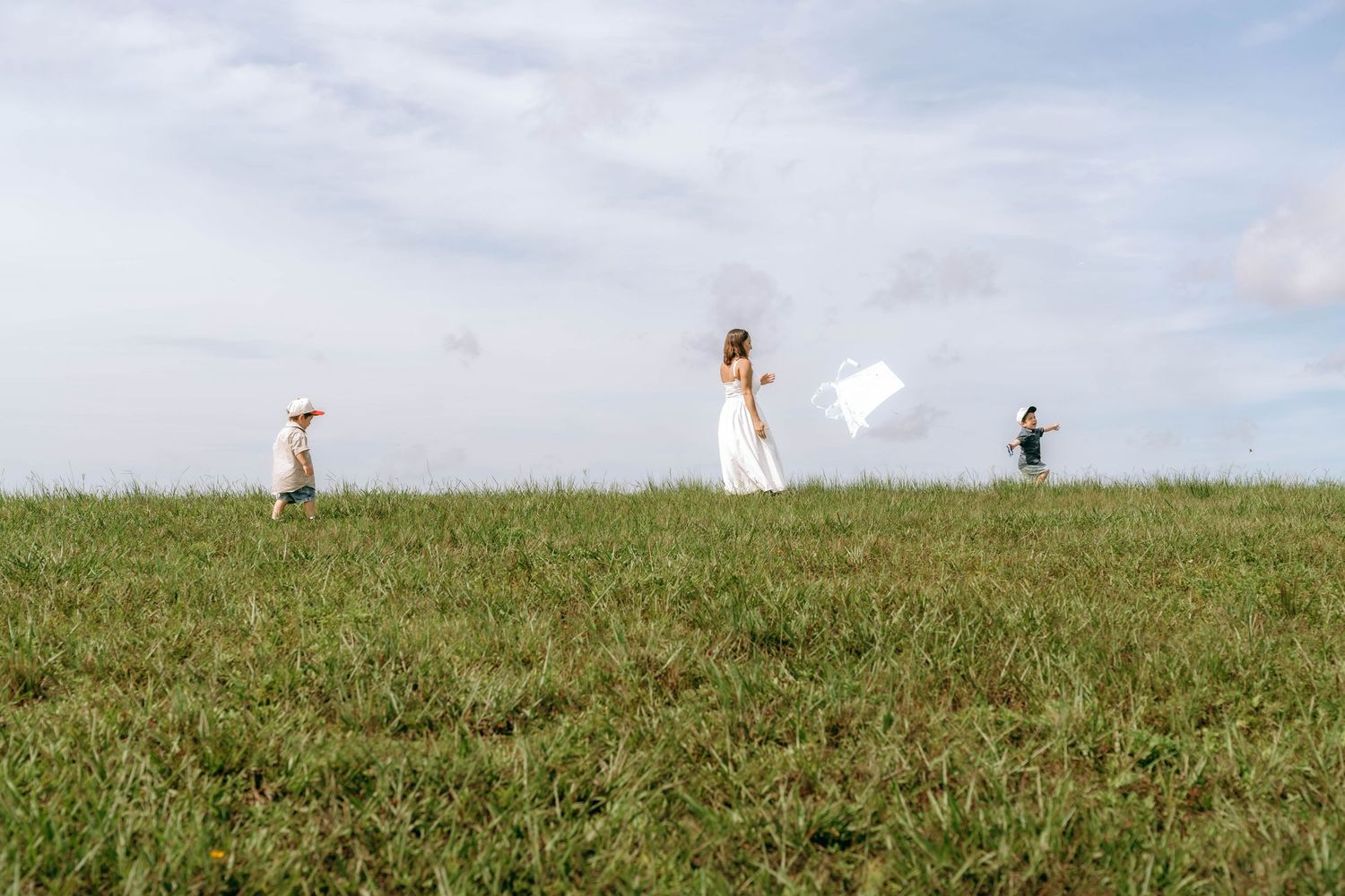 A woman in a white dress waves fabric in a green field while two children play nearby under a cloudy sky.