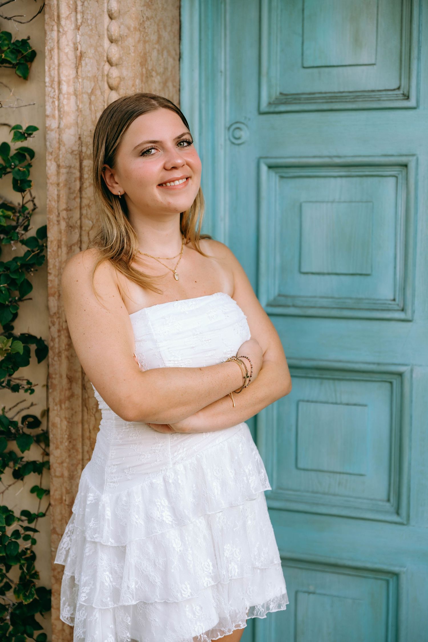 Young woman in white strapless ruffled dress smiling with arms crossed beside a teal vintage door.