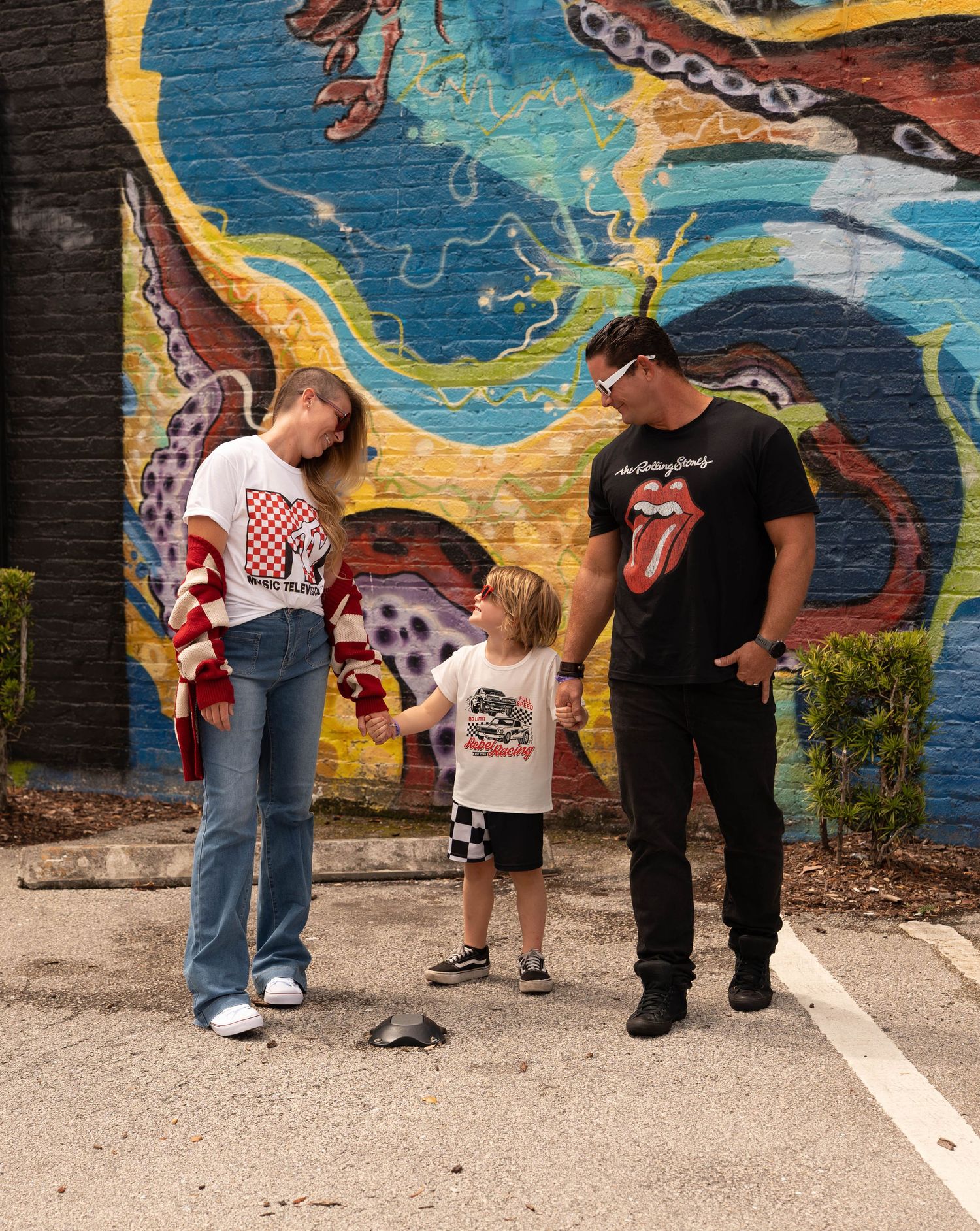 Family of three stands before a vibrant octopus mural, parents in Rolling Stones and checkered tees with toddler in Vans.