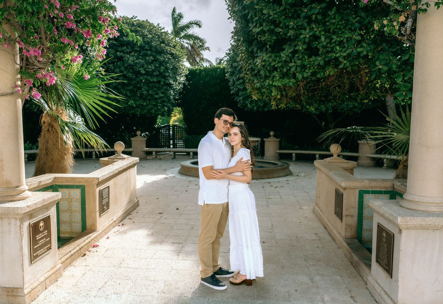 Couple embracing in a tropical garden courtyard with palm trees and pink flowers, wearing white outfits on Worth Avenue Palm beach.
