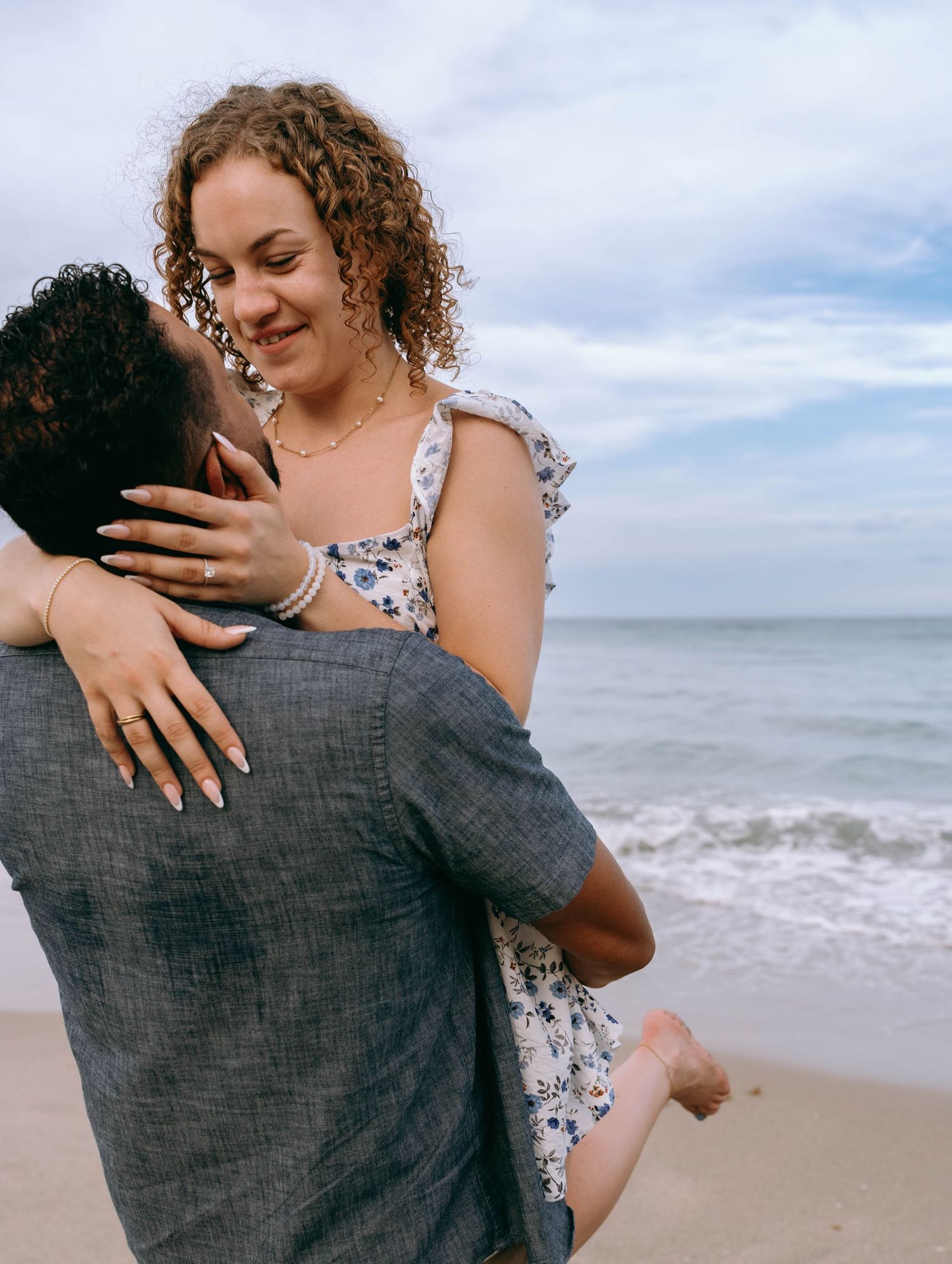 Man lifting smiling woman in floral dress on a beach, sharing a joyful romantic moment by the ocean on Worth Avenue Palm beach.