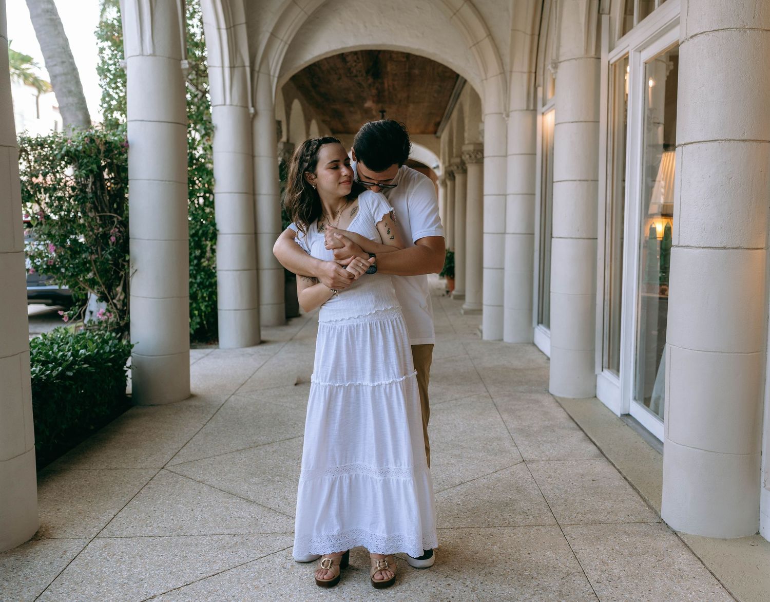 A couple embraces tenderly in a elegant columned walkway, the woman wearing a flowing white tiered maxi dress. Engagement photos Worth Ave