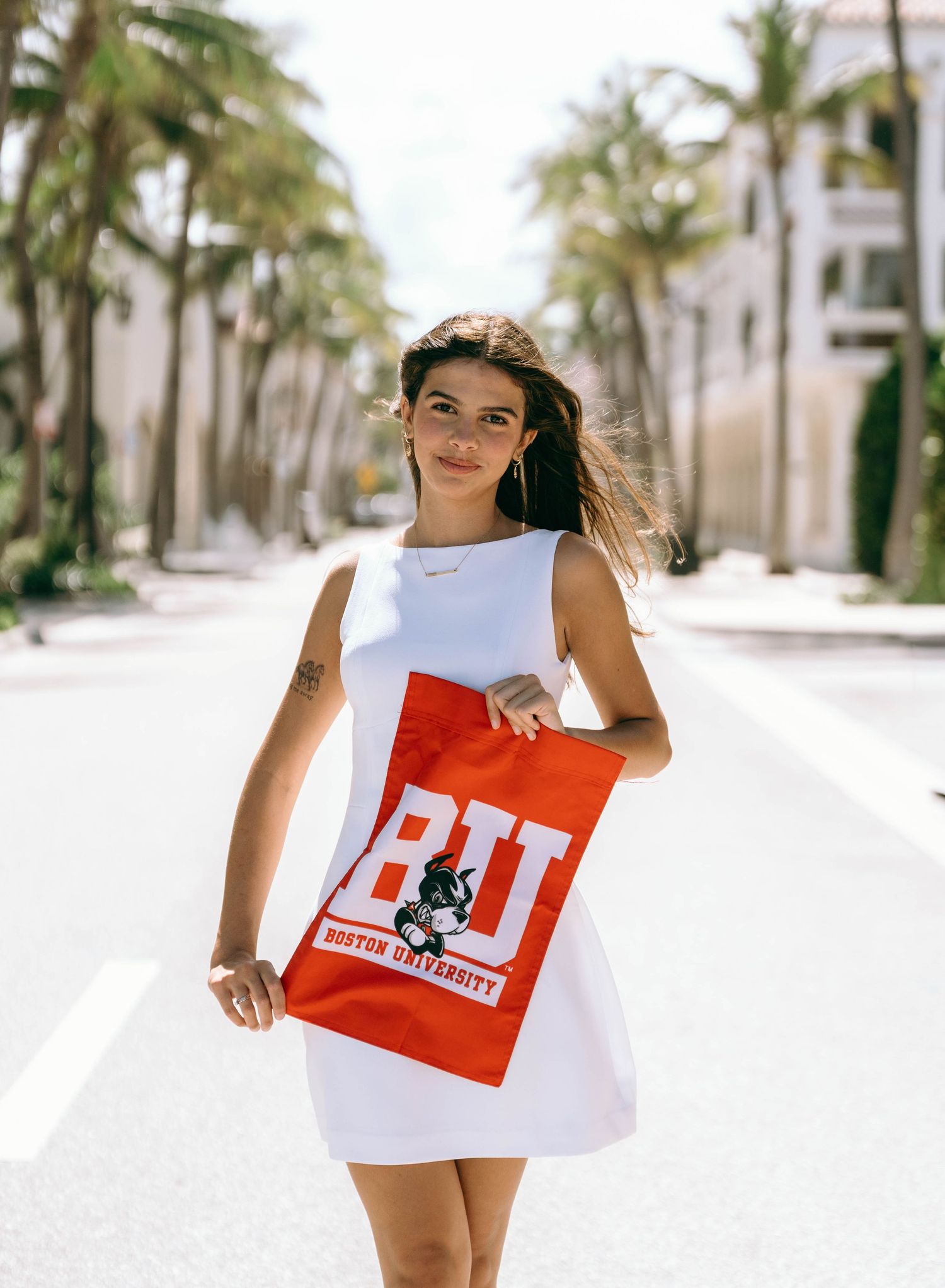Young woman in white dress holds Boston University red tote bag on sunny palm-lined street on Worth Avenue Palm beach.
