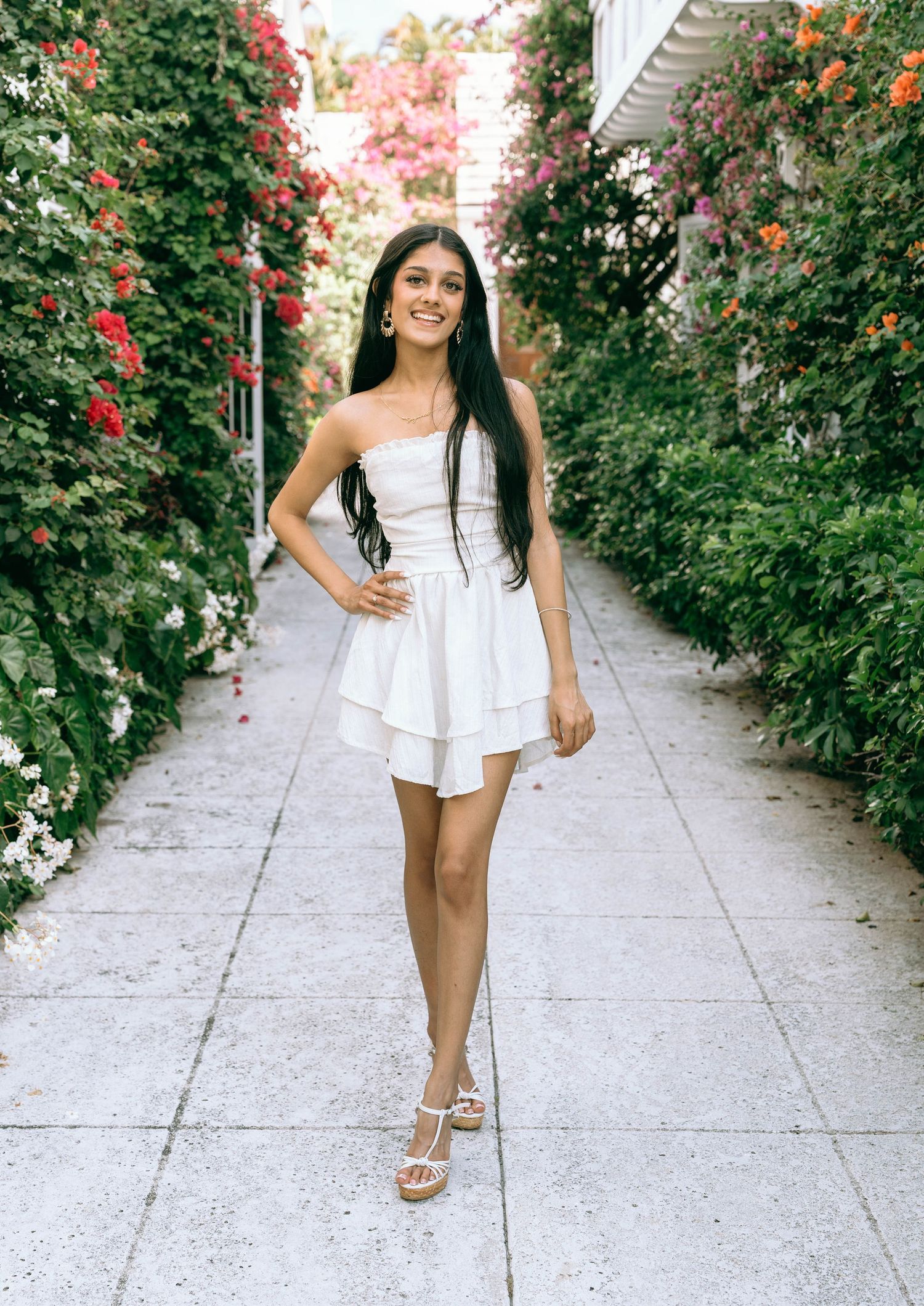 Smiling woman in white strapless mini dress walking through a vibrant floral garden corridor on Worth Avenue Palm beach.