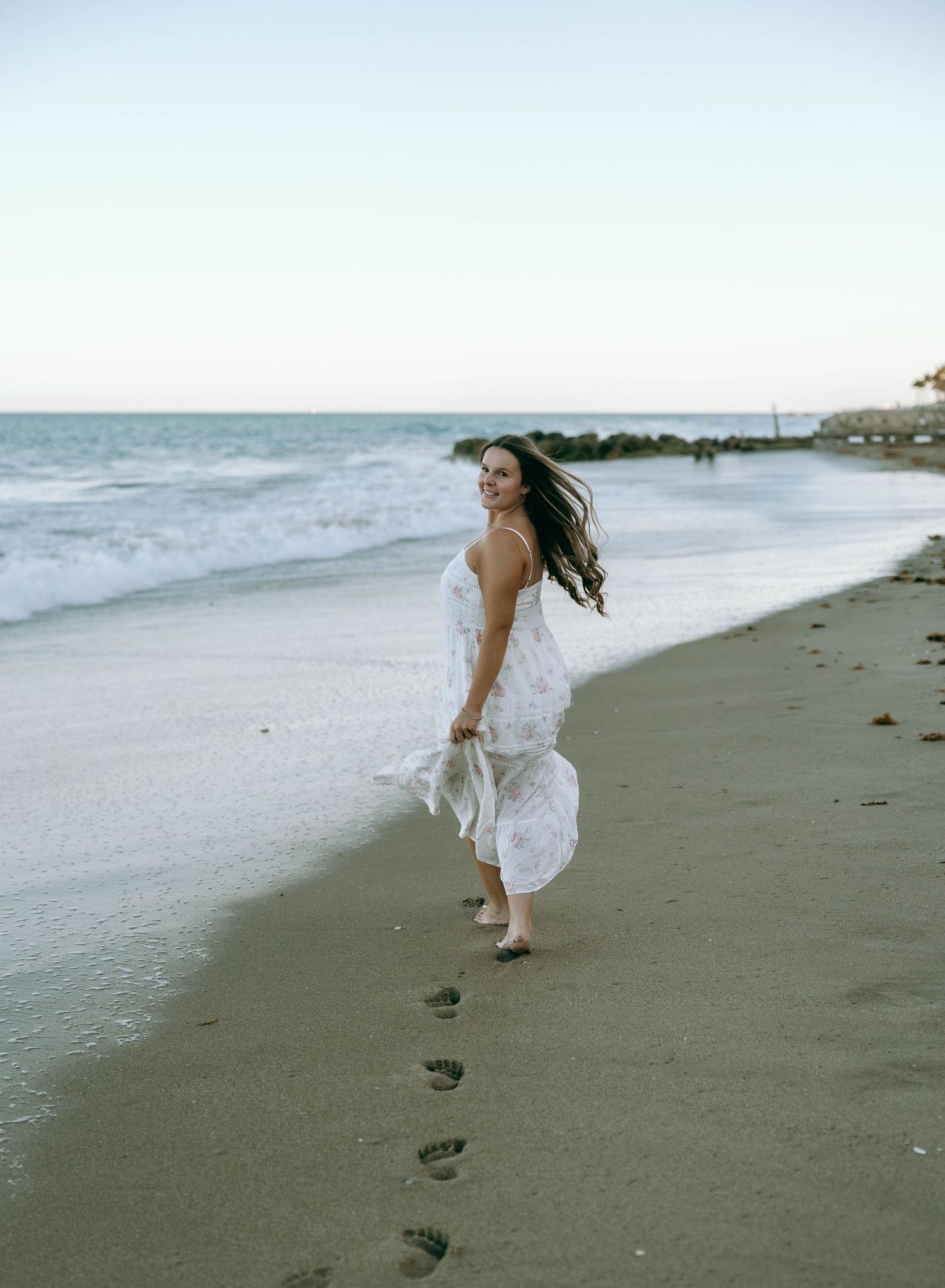 Woman in white maxi dress walking barefoot along sandy beach leaving footprints. Worth Avenue, Palm Beach, FL