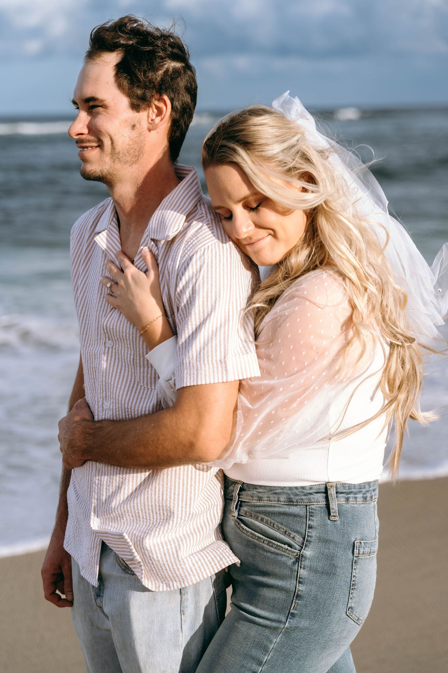 A couple laughs and plays together on a sunny beach with waves crashing behind them.