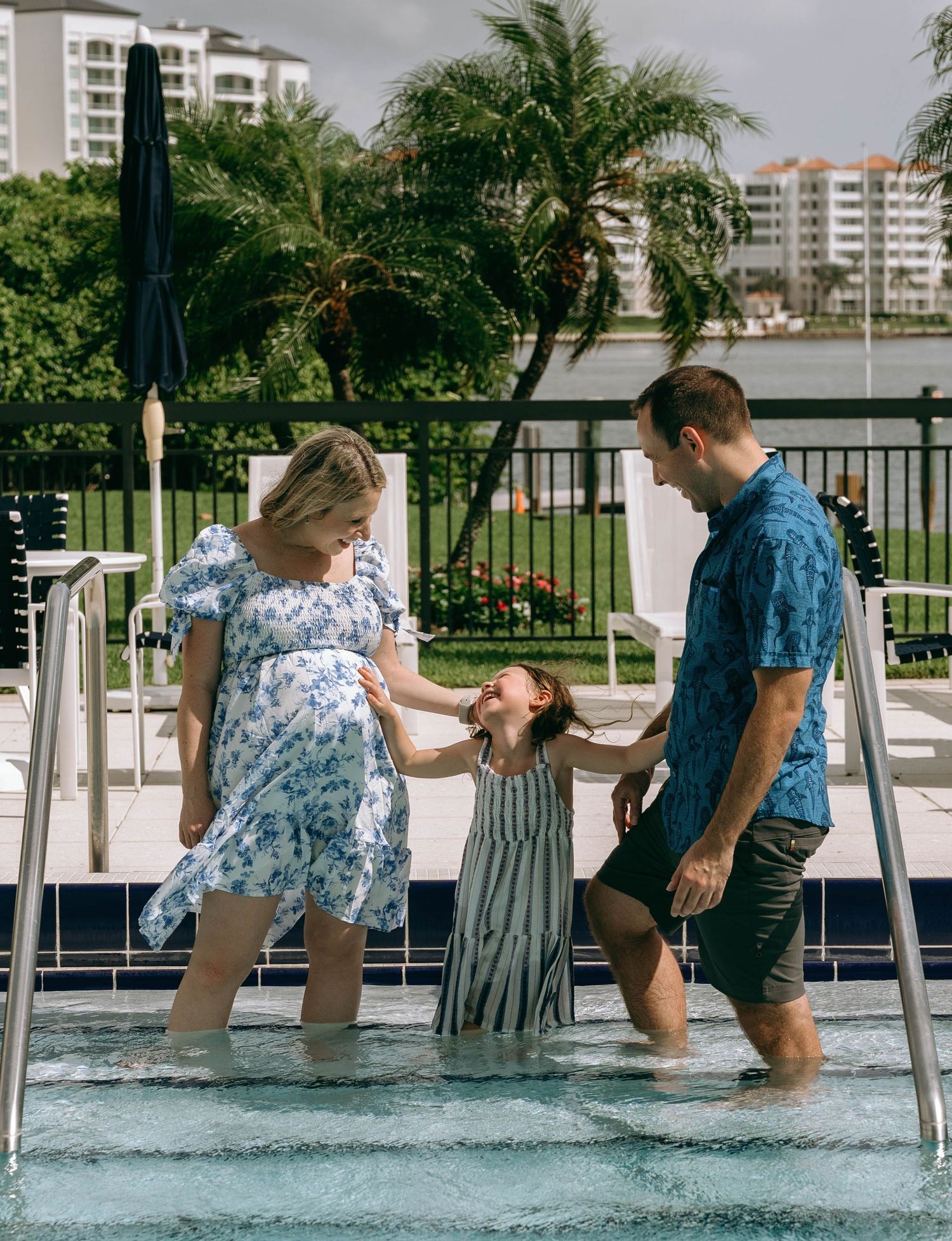 Pregnant woman in floral dress with husband and daughter playing in pool at tropical resort.