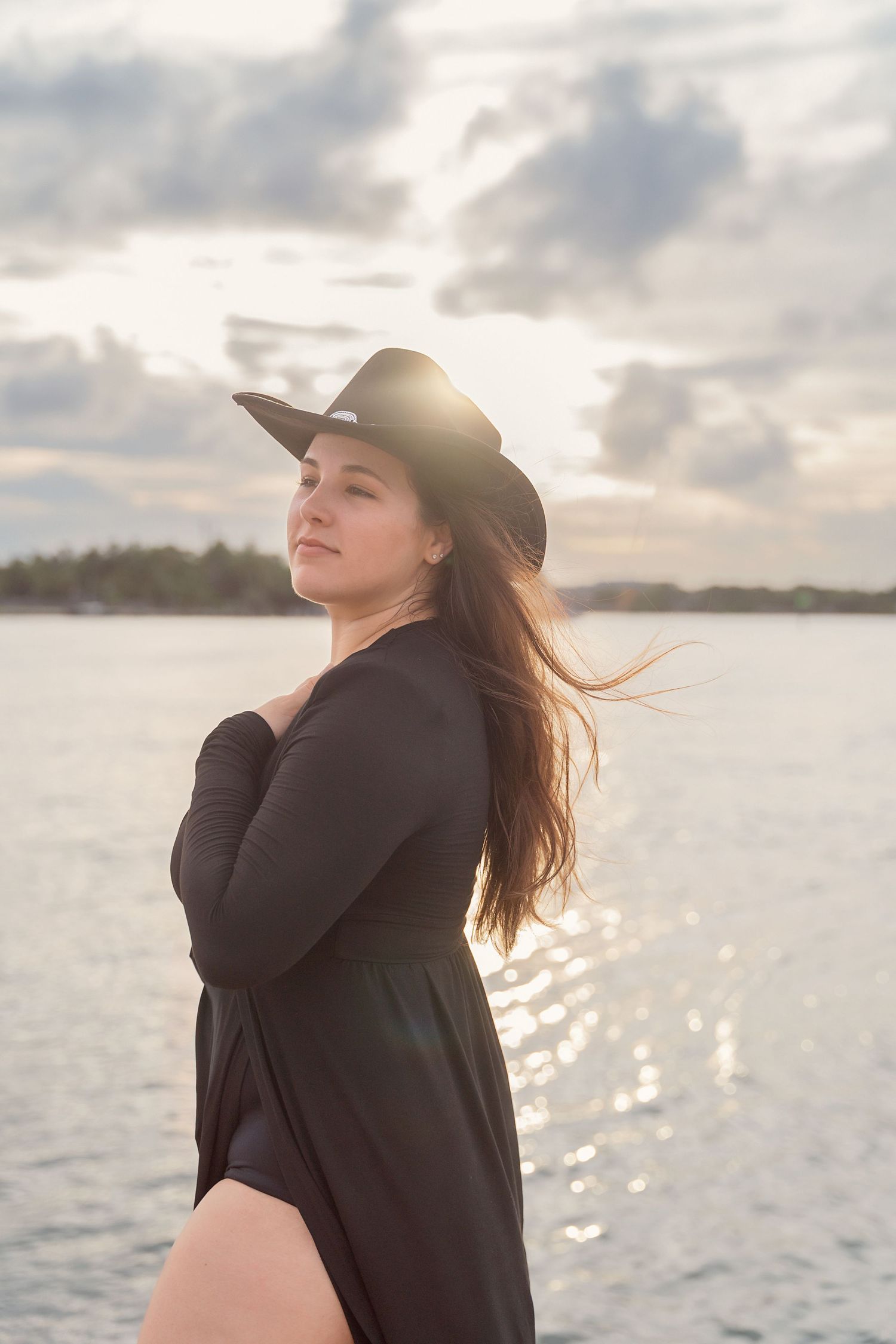 Woman in black dress and hat poses gracefully by the ocean during golden hour sunset.